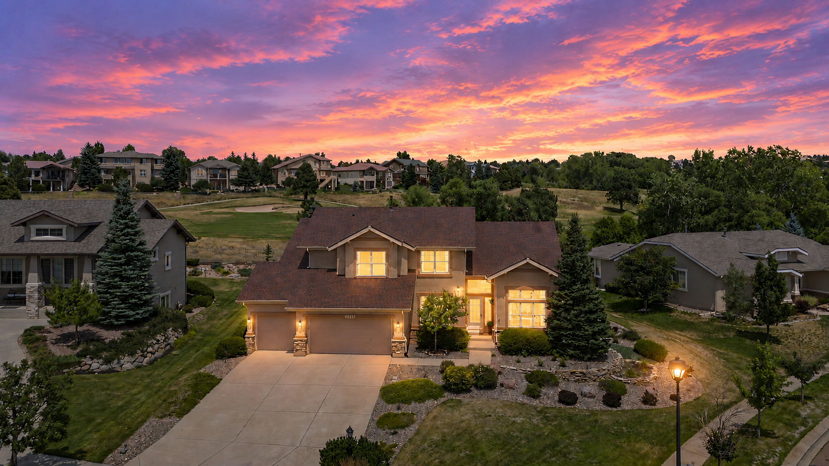 This elevated perspective captures a two-story suburban home at dusk, set against a dramatic, vibrant sunset sky. The residence features a multi-gabled roofline, a spacious concrete driveway leading to a multi-car garage, and a landscaped front yard with mature evergreens. The warm glow from the interior windows and exterior lighting creates an inviting atmosphere, highlighting the home's position overlooking a golf course.