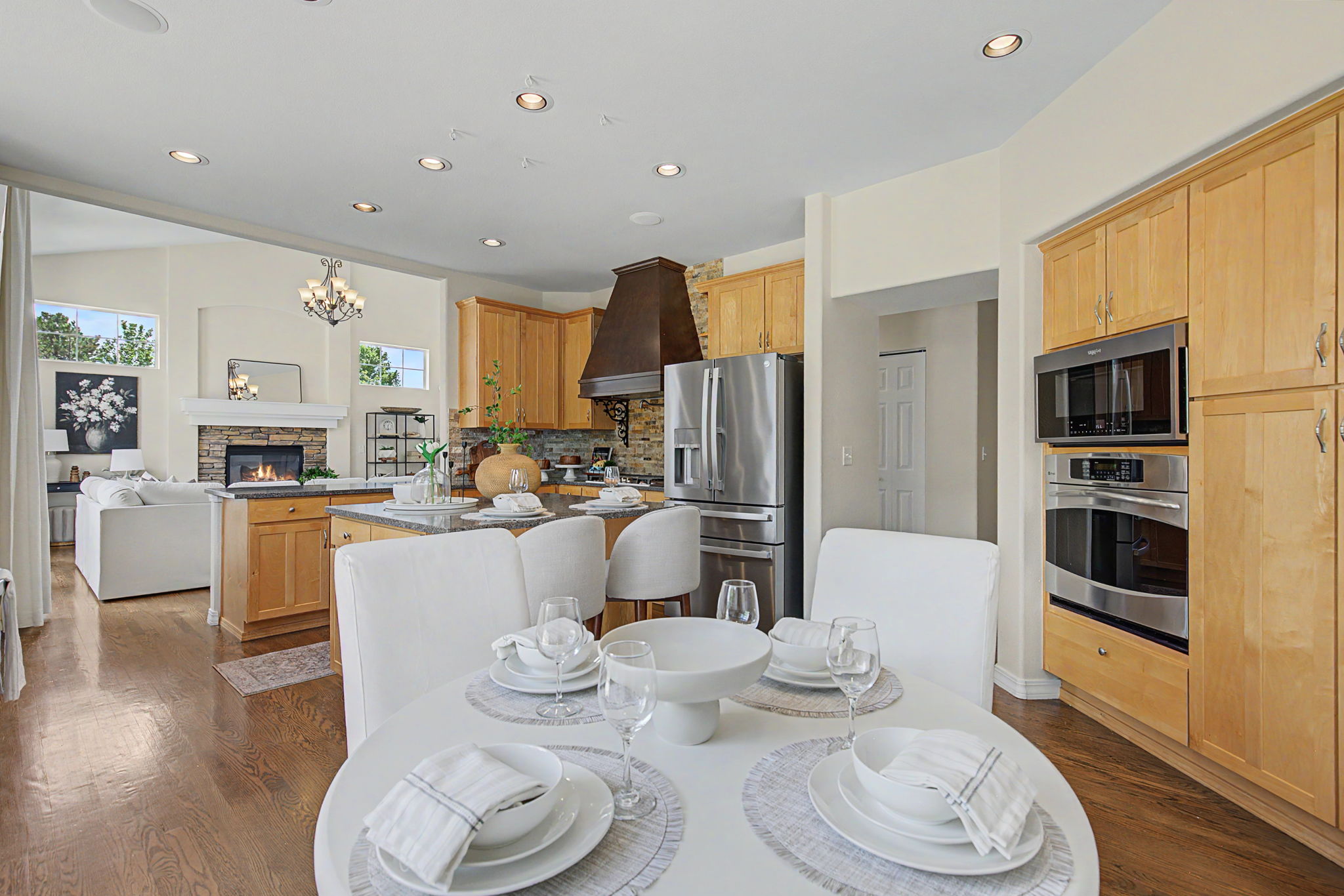 This open-concept kitchen and dining area features light wood cabinetry, stainless steel appliances, and a central island with stone countertops. A round dining table with white chairs sits in the foreground, while a cozy living area with a stone-clad fireplace is visible in the background. The space is bright and inviting, characterized by recessed lighting and a seamless flow between the cooking and living zones.