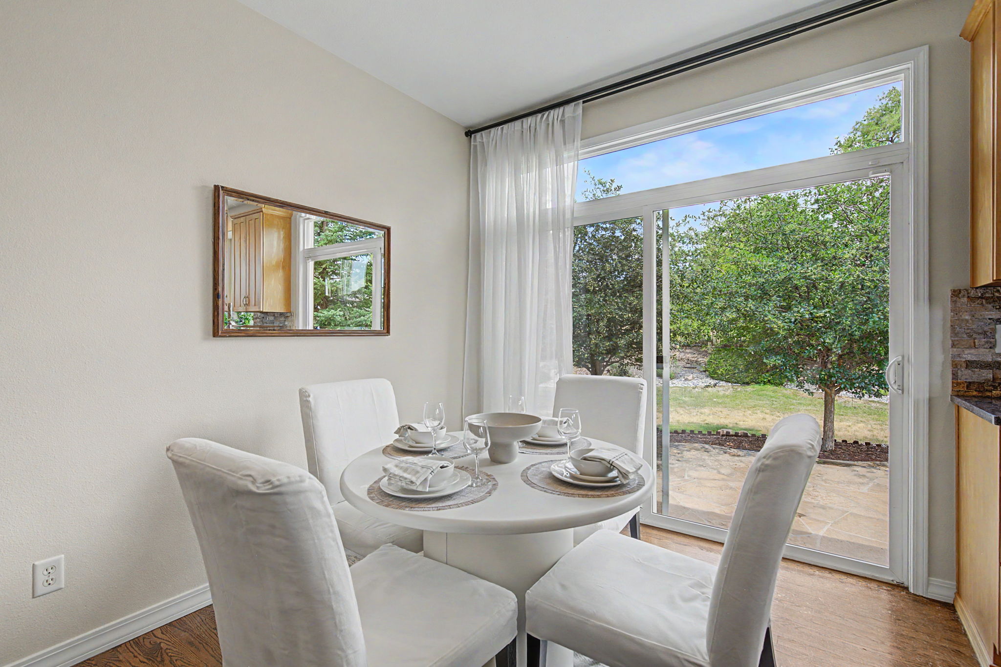 This bright dining area features a round white table set for four, surrounded by matching white upholstered chairs. A large sliding glass door provides abundant natural light and a clear view of the lush green backyard, while a rectangular mirror on the adjacent wall enhances the sense of space. The room is finished with warm wood-toned flooring, creating a clean and inviting atmosphere perfect for casual meals.