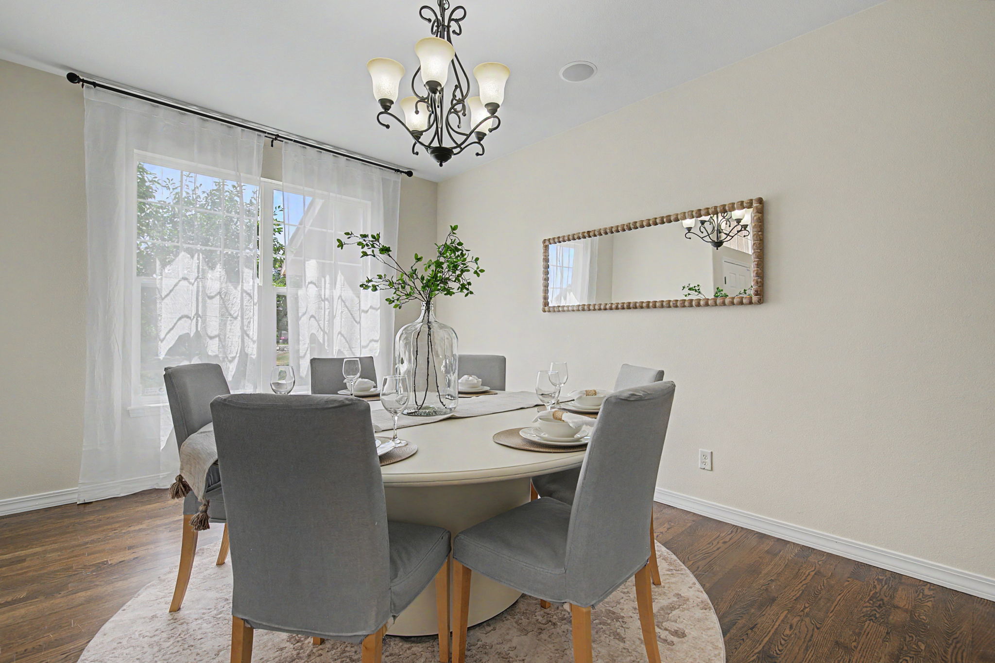 This elegant dining room features a round table surrounded by six upholstered grey chairs, set atop a circular patterned rug on dark wood flooring. A large glass vase with greenery serves as a centerpiece, while a decorative mirror hangs on the wall, reflecting the room and the ornate chandelier above. The space is brightened by large windows dressed in sheer white curtains, creating a welcoming and sophisticated atmosphere.