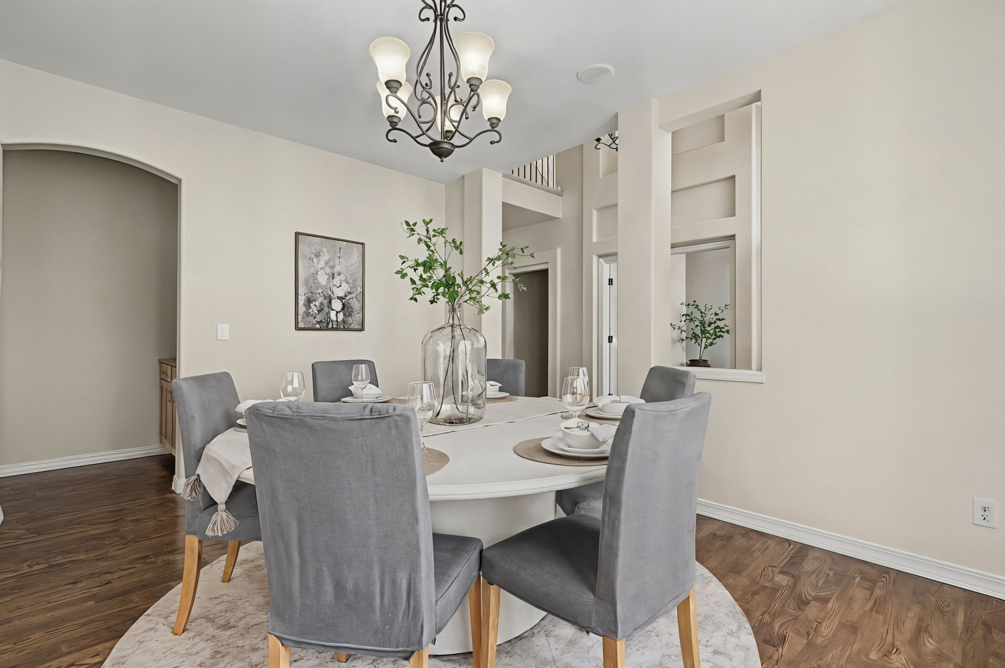This elegant dining room features a round white table surrounded by six grey upholstered chairs, set atop a circular patterned rug. A decorative chandelier hangs above the table, which is centered by a large glass vase filled with greenery. The room is finished with neutral walls, dark wood flooring, and architectural wall niches that add depth and character to the space.