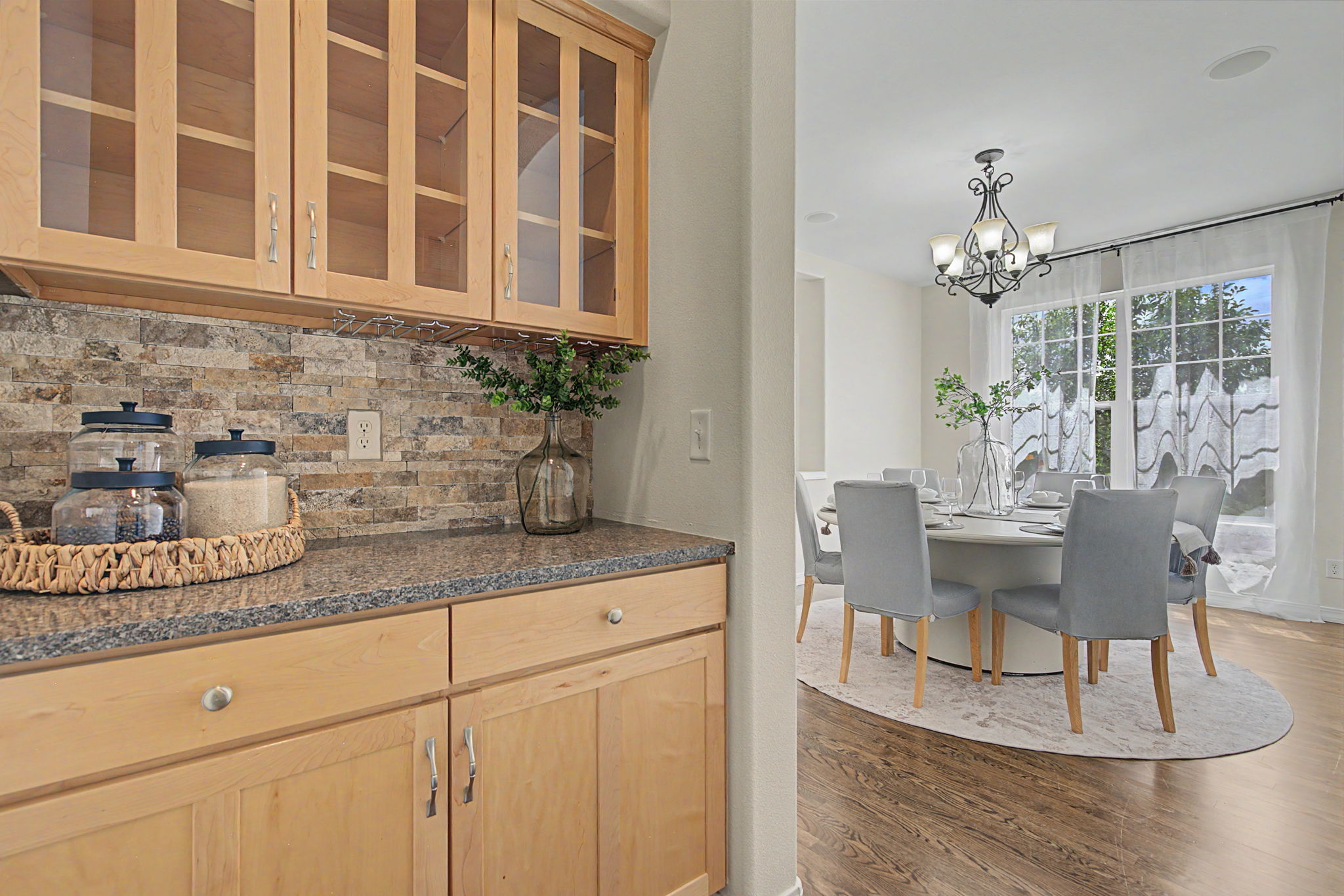 This image captures a transition between a kitchen butler's pantry and a formal dining room. The foreground features light wood cabinetry with glass-front upper doors, a stone-tiled backsplash, and a dark stone countertop holding glass storage jars. Through the doorway, a bright dining area is visible, furnished with a round table, grey upholstered chairs, and a decorative chandelier, all set against a backdrop of large windows draped in sheer curtains.