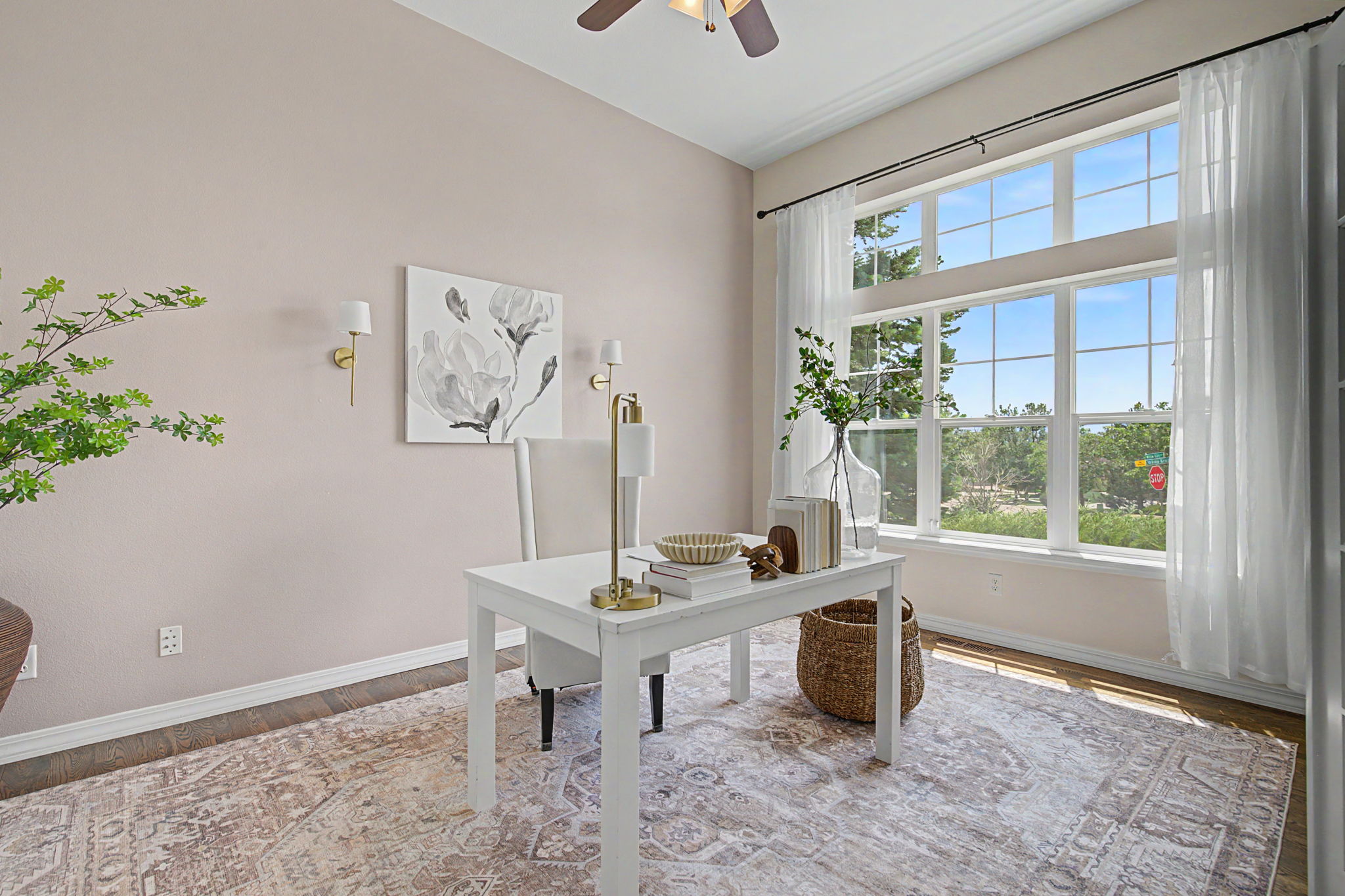 This bright and airy home office features soft, neutral-toned walls and a large, multi-pane window that floods the space with natural light. A minimalist white desk is centered on a large, patterned area rug, complemented by a high-backed chair and elegant gold-toned wall sconces. The room is accented with a decorative floral art piece and a large glass vase with greenery, creating a serene and professional atmosphere.