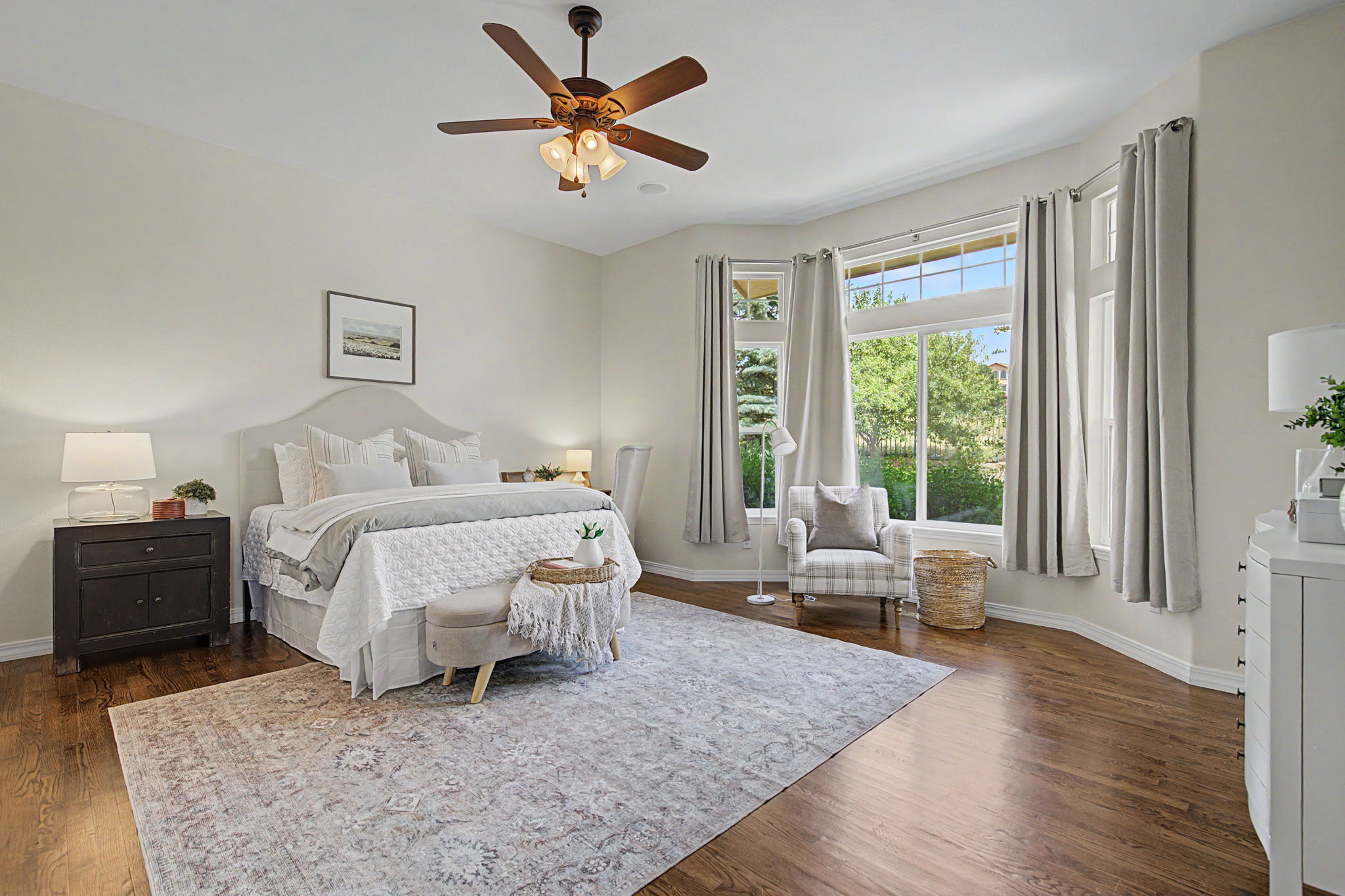 This spacious primary bedroom features a neutral color palette, hardwood flooring, and a large area rug that anchors the space. A king-sized bed with layered bedding sits against a light-colored wall, flanked by a dark wood nightstand and a matching lamp. Large windows with soft curtains allow natural light to fill the room, highlighting a cozy seating area with a plaid armchair and a woven basket.