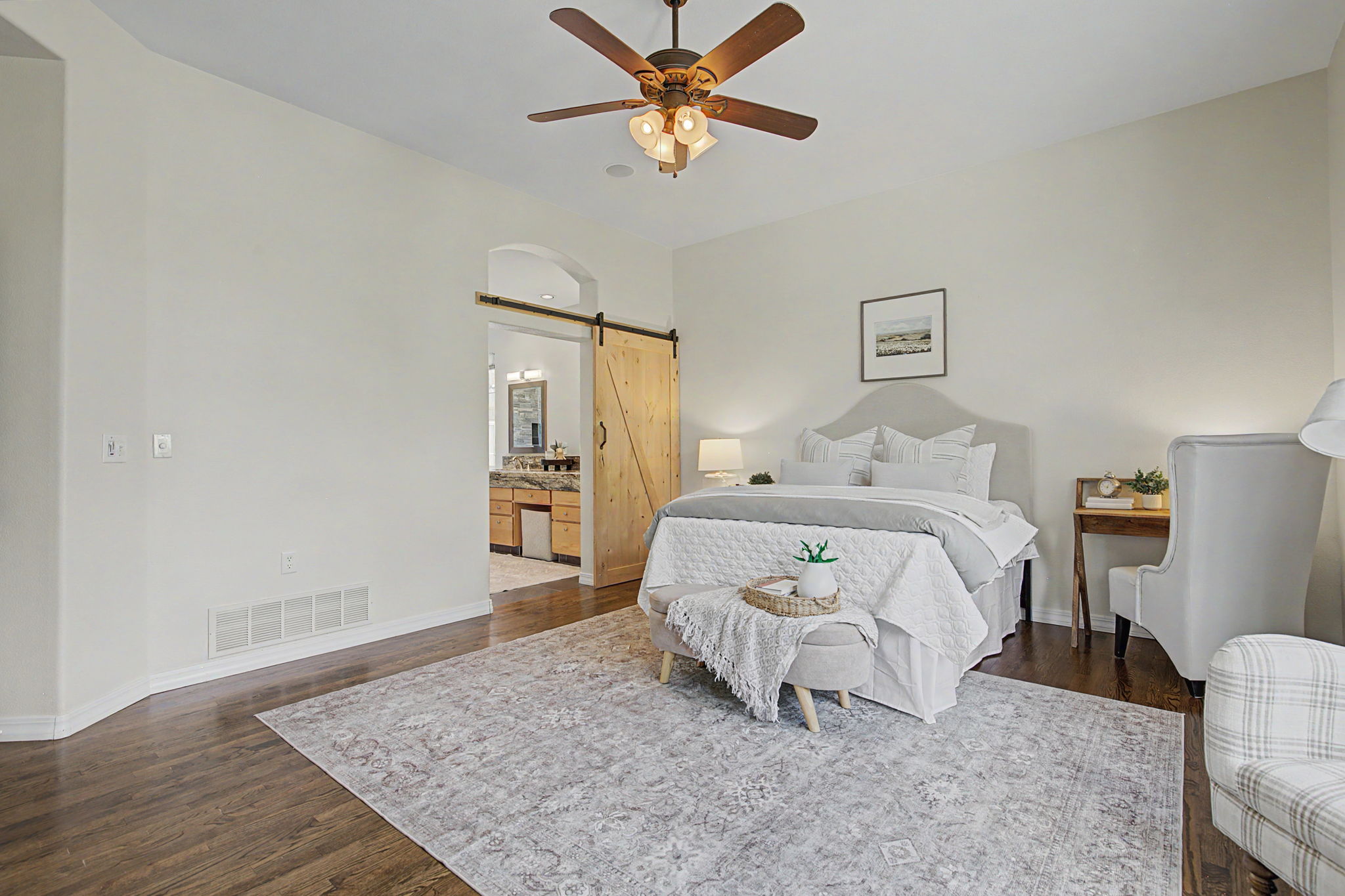 This primary bedroom features a neutral color palette with a plush upholstered headboard and a patterned area rug over dark wood flooring. A sliding barn door provides access to an adjoining bathroom, while a small desk area and a ceiling fan with integrated lighting complete the space. The room is captured from a wide-angle perspective, highlighting the balanced layout and inviting, serene atmosphere.