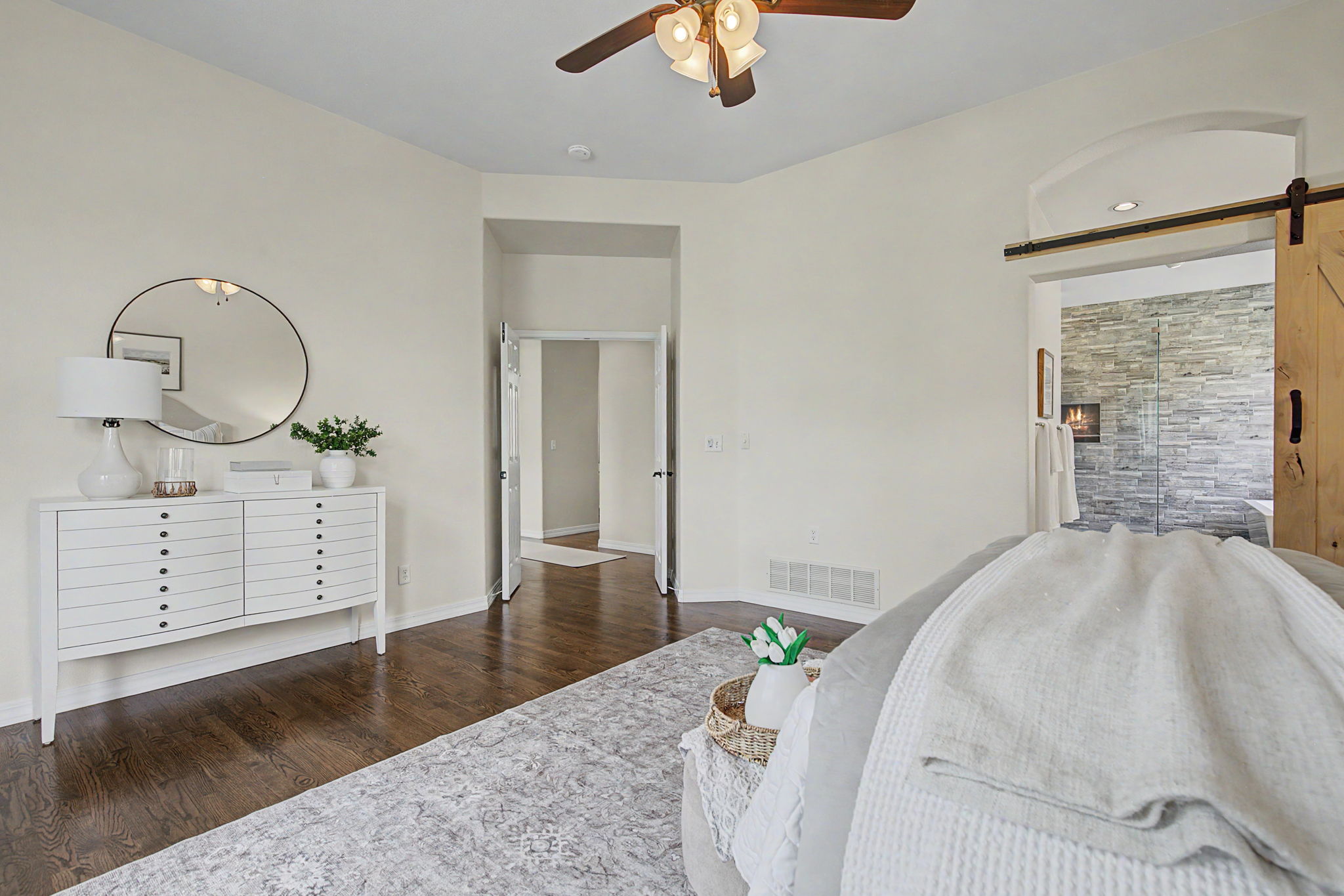 This primary bedroom features a neutral color palette with dark hardwood floors and a large, patterned area rug. A white dresser with a round mirror sits against the wall, while a sliding barn door leads to an adjacent space with a stone-clad wall and a visible fireplace. The room is illuminated by a ceiling fan with integrated lighting, creating a bright and inviting atmosphere.
