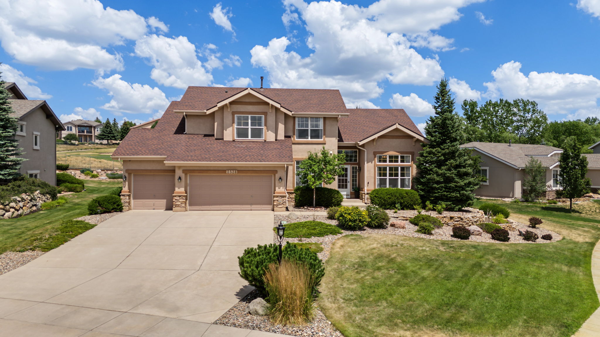 This elevated front-facing view captures a two-story suburban home with a tan stucco facade and a multi-gabled brown shingle roof. The property features a spacious concrete driveway leading to a three-car garage, complemented by mature landscaping, a prominent evergreen tree, and a well-maintained lawn. The bright blue sky with scattered clouds provides a welcoming backdrop for this traditional residential architecture.