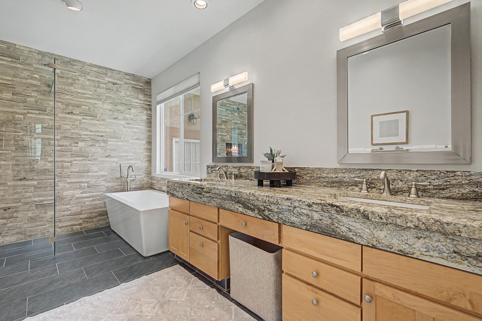This primary bathroom features a dual-sink vanity with a stone countertop and light wood cabinetry, complemented by two rectangular mirrors and wall-mounted lighting. To the left, a freestanding soaking tub is positioned against a wall finished with textured stone tiles, adjacent to a glass-enclosed shower area. The space is grounded by dark, rectangular floor tiles and a light-colored area rug, creating a clean and functional aesthetic.