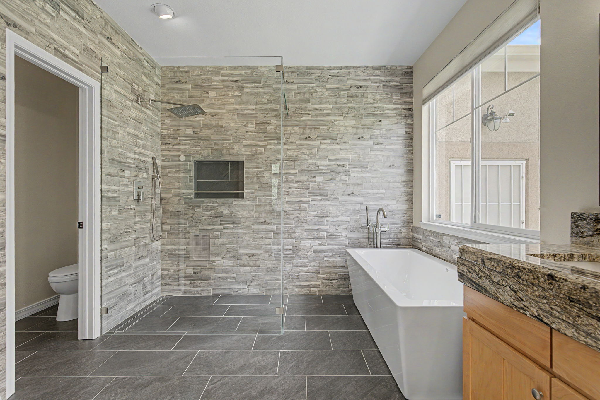 This modern primary bathroom features a spacious walk-in shower with a glass enclosure and a separate freestanding soaking tub. The walls are adorned with textured, horizontal stone tiles, complemented by large-format dark gray floor tiles. A large window above the tub provides natural light, while a wooden vanity with a stone countertop adds a warm, organic touch to the space.