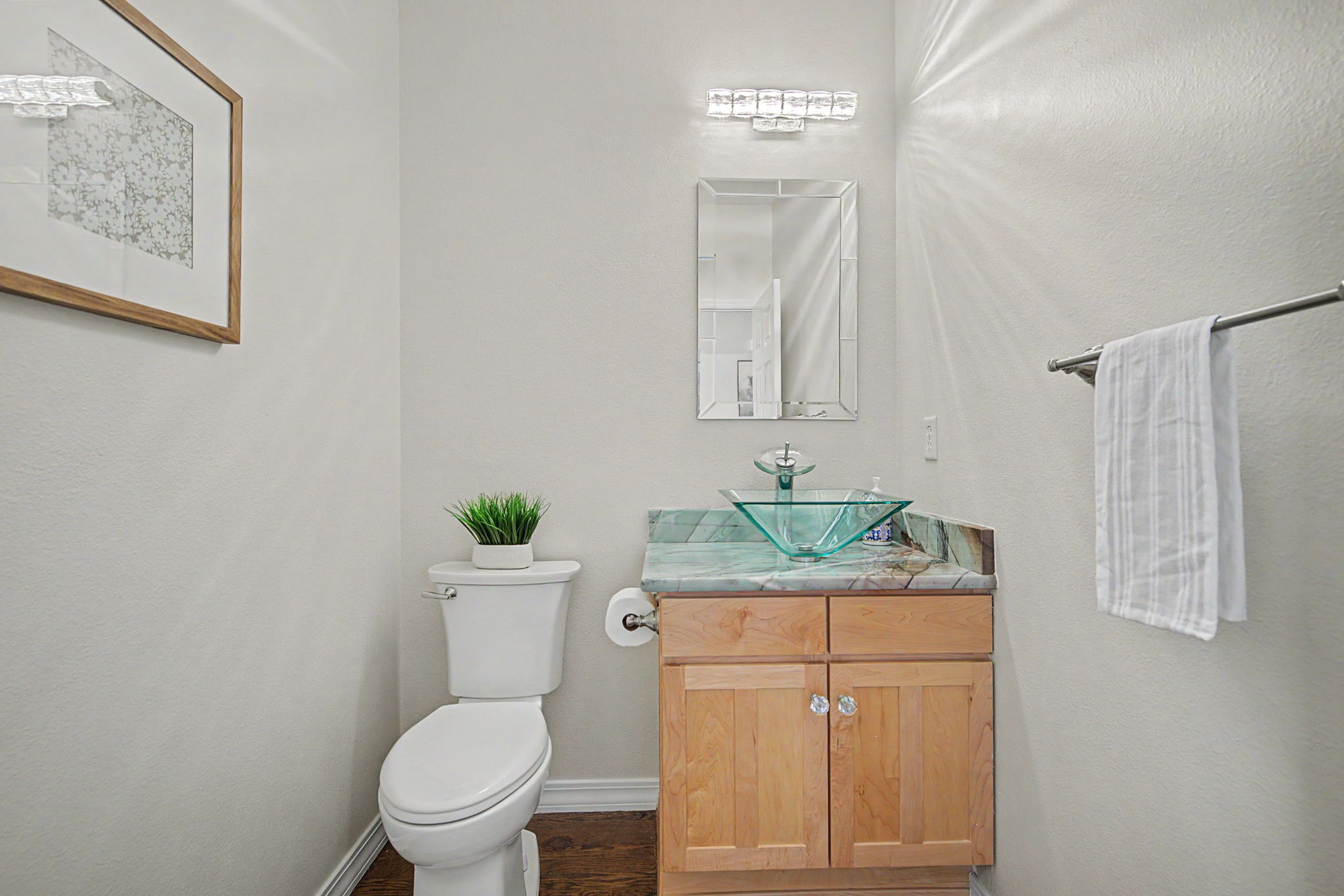 This compact powder room features a light wood vanity topped with a green-toned stone counter and a unique, square glass vessel sink. A rectangular mirror is centered above the vanity, illuminated by a modern multi-light fixture, while a white toilet sits to the left. The space is finished with neutral walls, a framed piece of art, and a towel bar, creating a clean and functional aesthetic.
