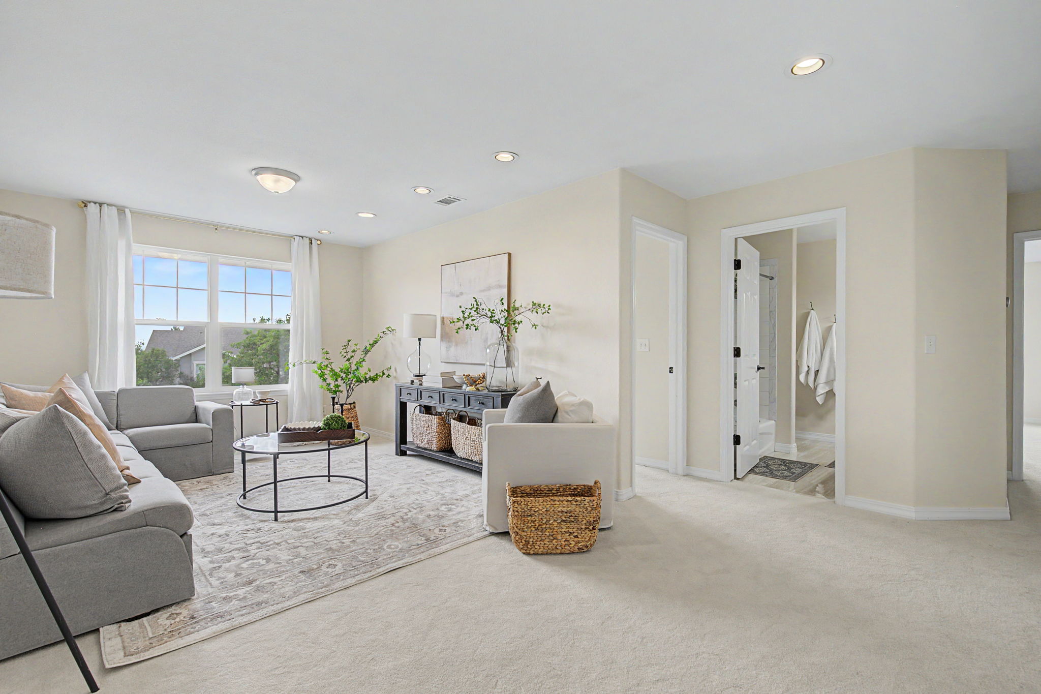 This bright and inviting living room features a neutral color palette with soft gray seating and a patterned area rug. A dark console table with woven storage baskets sits behind a white armchair, while a glass-topped coffee table centers the space. The room is illuminated by recessed lighting and natural light from large windows, with a doorway leading to an adjacent bathroom.