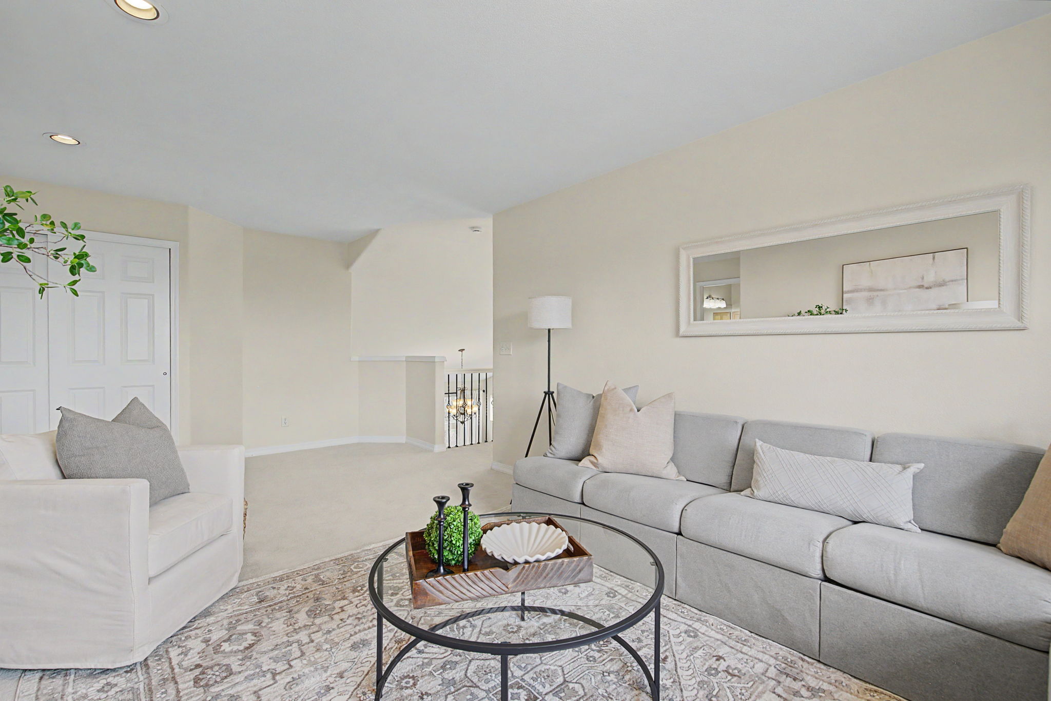 This bright and inviting living room features a neutral color palette with a light gray sofa and a matching armchair, accented by soft-toned throw pillows. A round glass-topped coffee table with a decorative wooden tray sits atop a patterned area rug, while a large rectangular mirror hangs above the sofa to enhance the sense of space. The room is illuminated by recessed lighting and a sleek floor lamp, creating a clean, modern, and comfortable atmosphere.