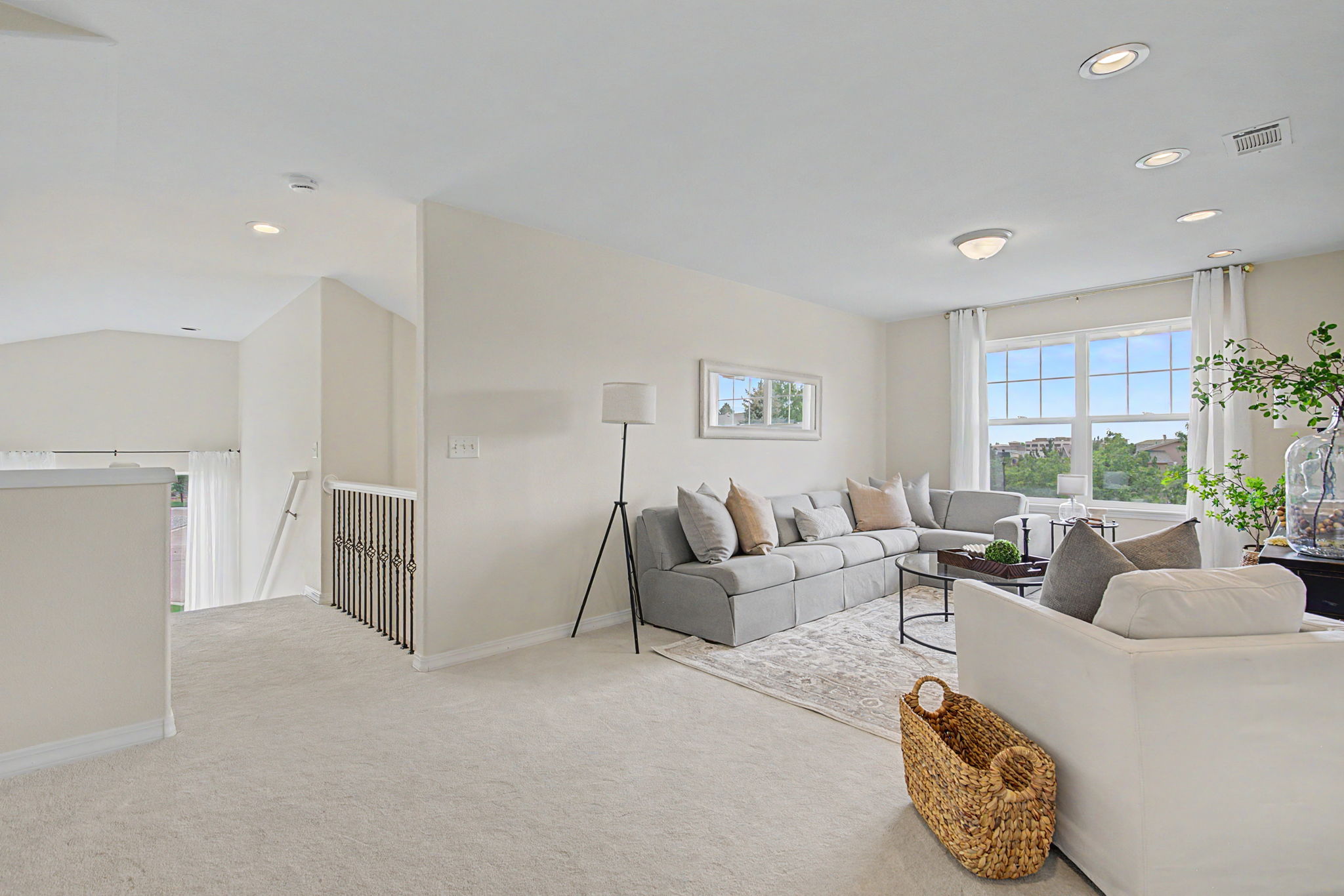 This bright and airy living room features a neutral color palette with a large sectional sofa arranged on a patterned area rug. The space is illuminated by recessed lighting and natural light streaming through large windows, while a sleek floor lamp and a woven basket add texture to the room. The open-concept layout connects to a staircase area with decorative metal railings, creating a spacious and inviting atmosphere.