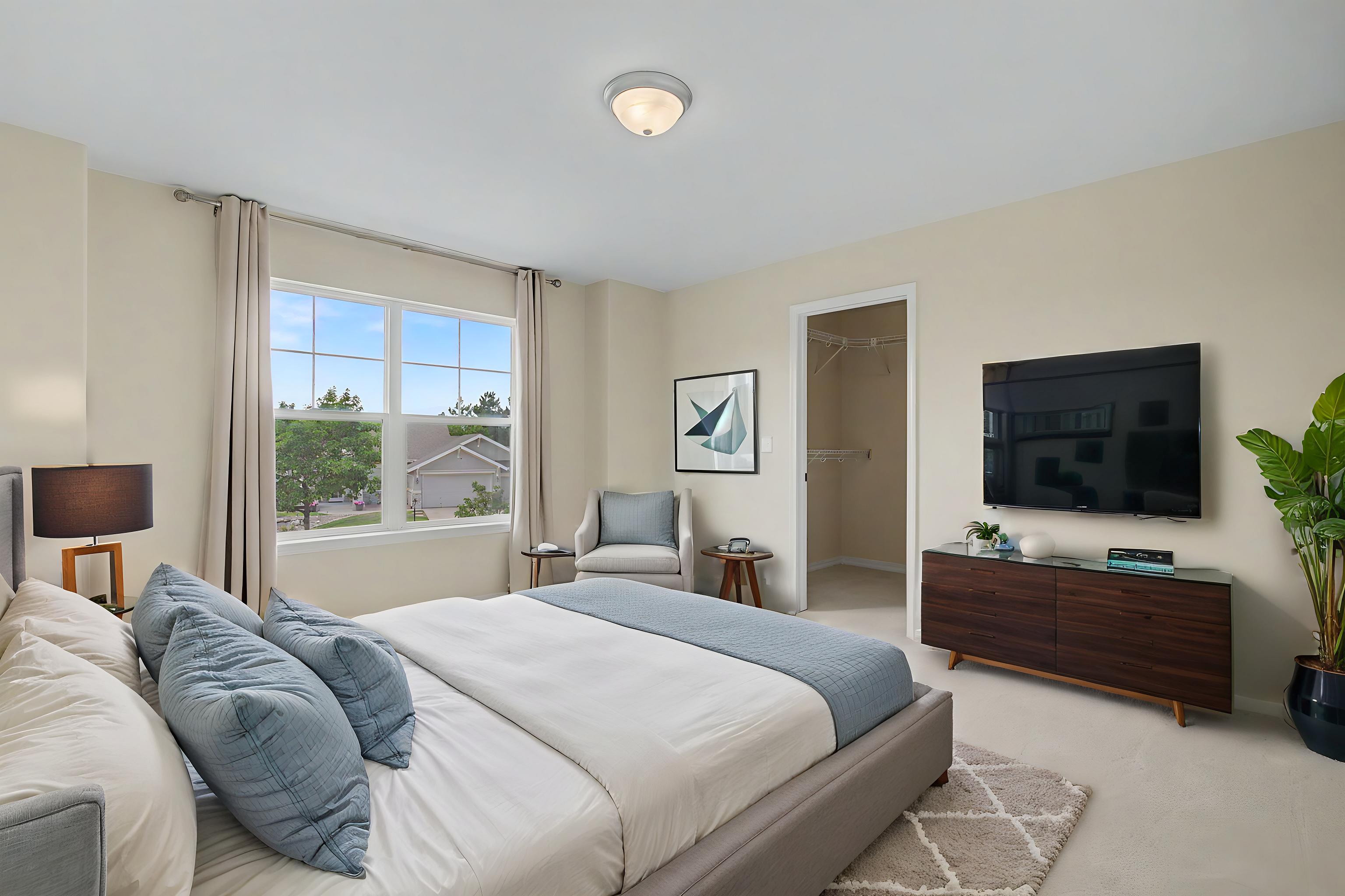 This primary bedroom features a neutral color palette with soft blue accents, centered around a plush upholstered bed. A dark wood dresser sits beneath a wall-mounted television, while a cozy armchair and side table are positioned near a large window that provides natural light and a view of the neighborhood. The room includes a walk-in closet entrance and a potted plant, creating a serene and well-appointed living space.