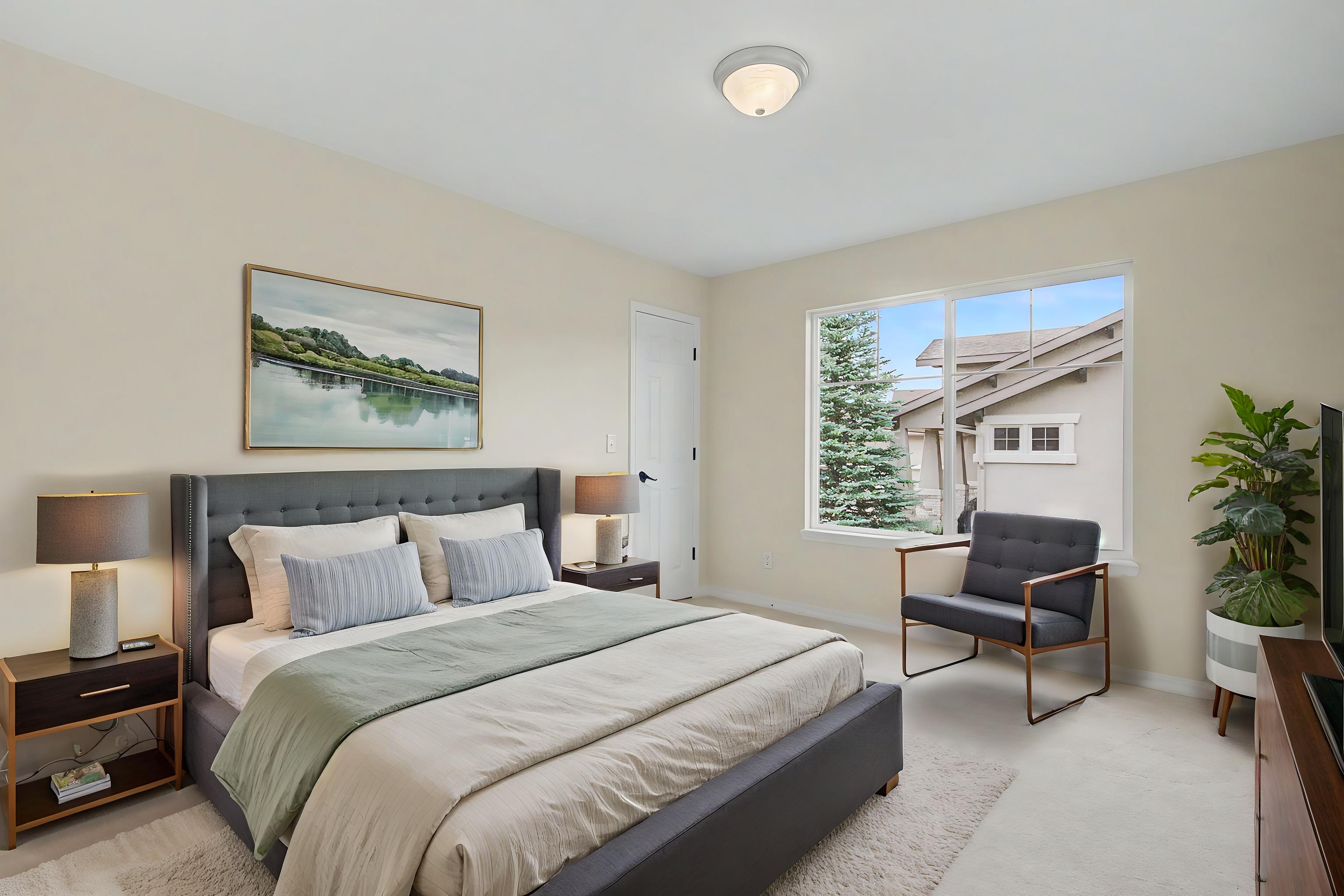 This primary bedroom features a contemporary gray upholstered bed frame as the focal point, complemented by a neutral color palette and soft lighting. A large window allows natural light to fill the room, highlighting the minimalist decor, a modern armchair, and a potted plant in the corner. The space is well-appointed with matching bedside tables and lamps, creating a serene and inviting atmosphere.