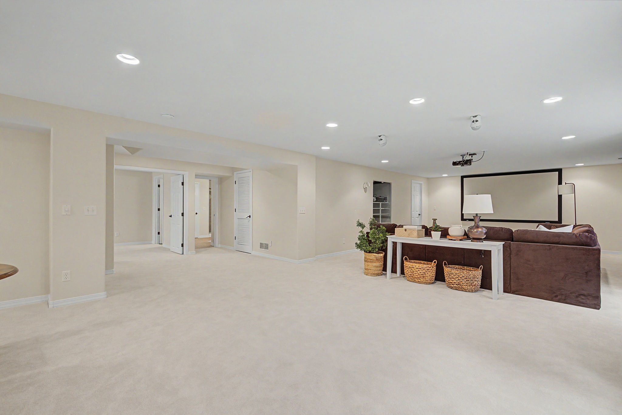 This spacious, neutral-toned basement features plush carpeting and recessed lighting throughout, creating a versatile living area. A large brown sectional sofa is positioned in front of a projector screen, complemented by a white console table with woven baskets and a table lamp. The open floor plan leads to multiple doorways, suggesting access to additional rooms or storage areas.