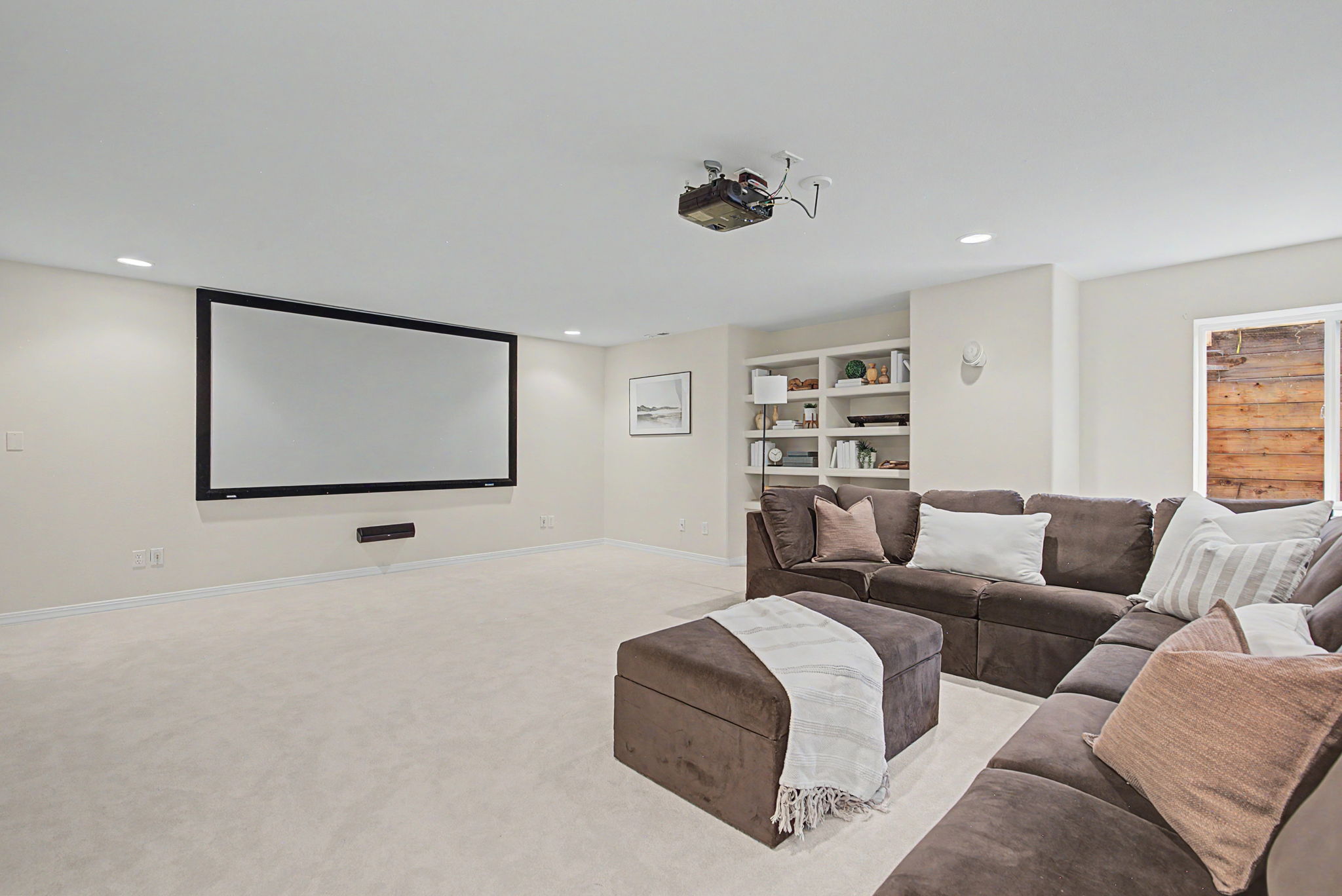 This spacious basement media room features a large projector screen mounted on a neutral wall, complemented by a ceiling-mounted projector. A plush, dark brown sectional sofa with an accompanying ottoman provides comfortable seating, while built-in shelving units offer display space for decor. The room is finished with light-colored carpeting and recessed lighting, creating a cozy and functional entertainment area.