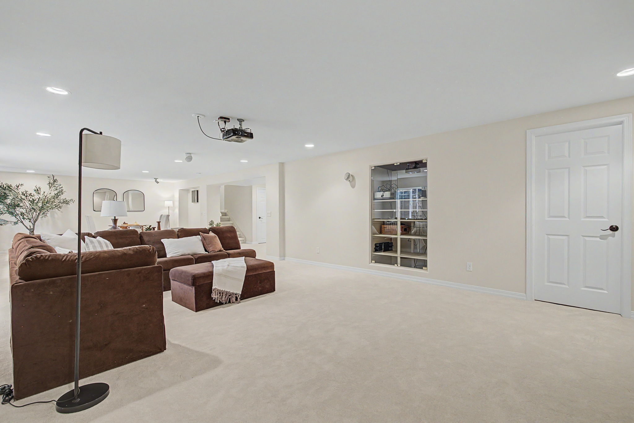 This spacious, carpeted basement features a large brown sectional sofa and a matching ottoman, creating a comfortable lounge area. A ceiling-mounted projector is positioned to face the opposite wall, which includes a built-in glass-fronted display cabinet. The room is finished with neutral-toned walls and recessed lighting, offering a versatile space for entertainment or relaxation.