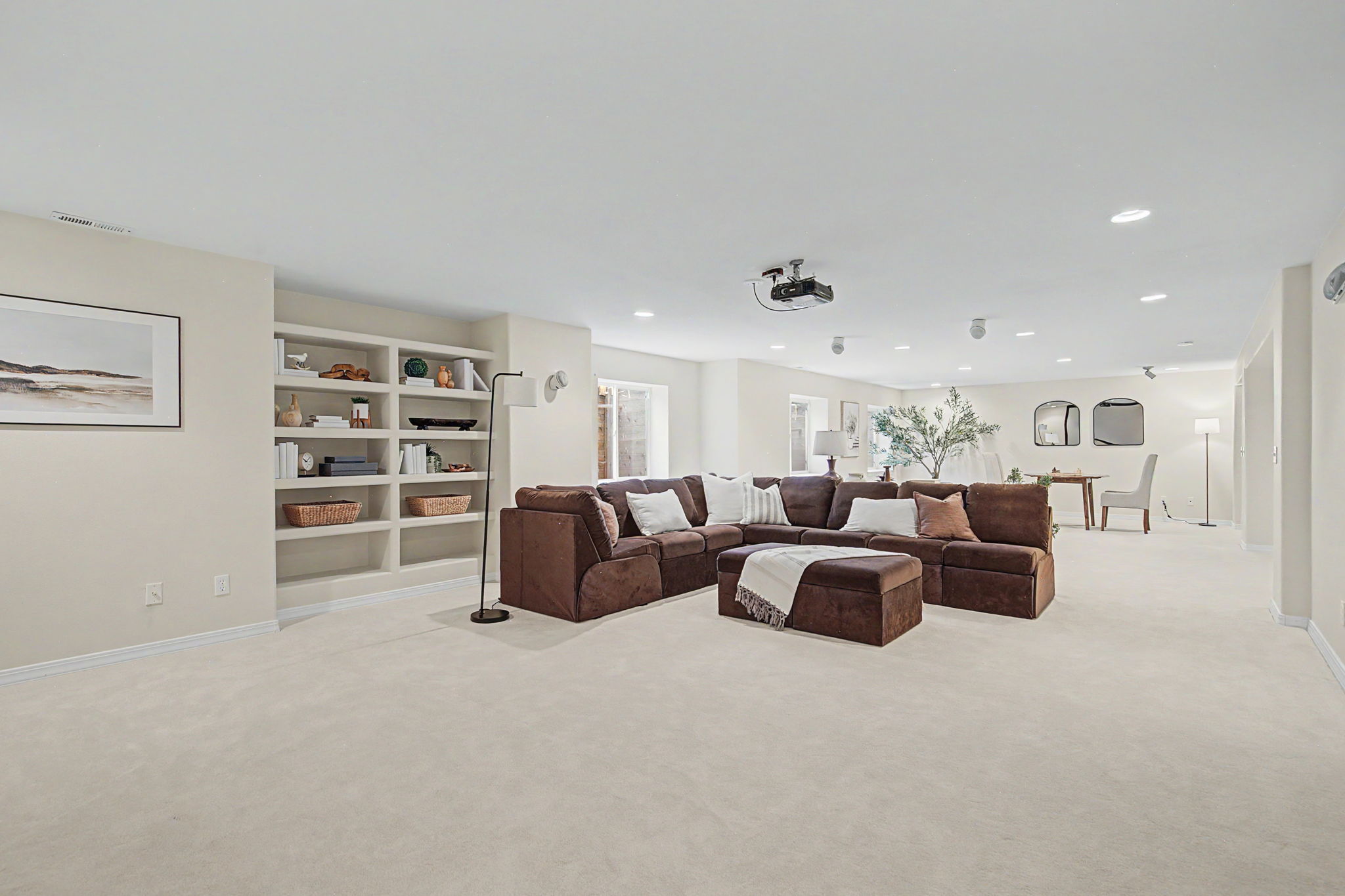 This spacious, open-concept basement features a large, plush brown sectional sofa arranged around a matching ottoman, creating a comfortable lounge area. Built-in shelving units provide ample storage and display space, while a projector mounted to the ceiling suggests a dedicated home theater setup. The room is finished with neutral carpeting and recessed lighting, offering a versatile and inviting space for relaxation or entertainment.