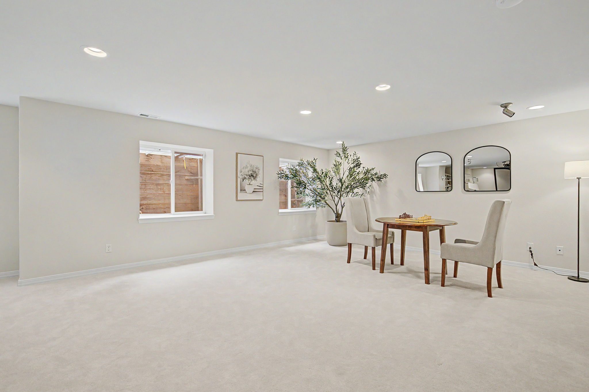 This spacious, neutrally-toned basement features plush carpeting and recessed lighting, creating a clean and versatile living area. A small dining set with two upholstered chairs sits near a large potted plant, while two arched mirrors add a decorative touch to the wall. The room is illuminated by natural light from two windows, offering a bright and inviting atmosphere suitable for a variety of uses.