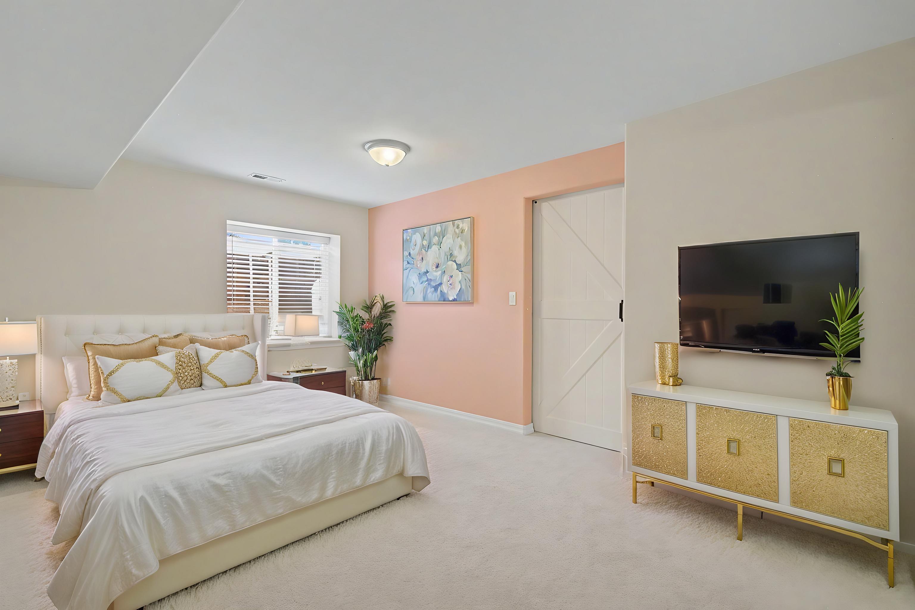 This primary bedroom features a neutral color palette accented by a soft pink feature wall and gold-toned furniture accents. A plush white bed with decorative pillows serves as the focal point, complemented by a modern white and gold media console holding a wall-mounted television. The room is brightened by natural light from a window and includes a sliding barn-style door, creating a contemporary and inviting atmosphere.