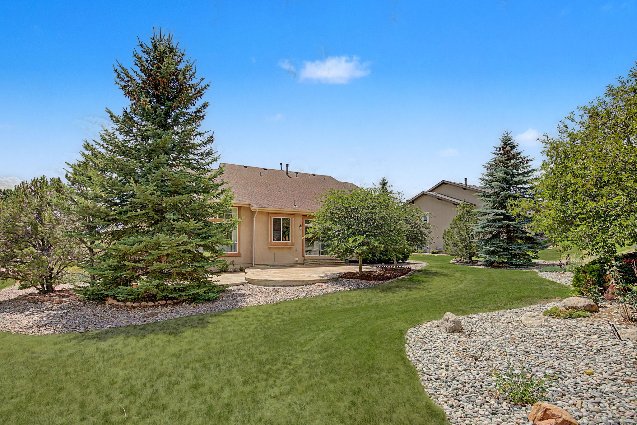 This rear view of a suburban home showcases a well-maintained backyard featuring a curved lawn bordered by decorative rock landscaping. A stone patio extends from the back of the house, providing a functional outdoor living space near a mature tree and large evergreen. The scene is captured from a ground-level perspective, highlighting the open, landscaped grounds and the home's neutral-toned exterior.