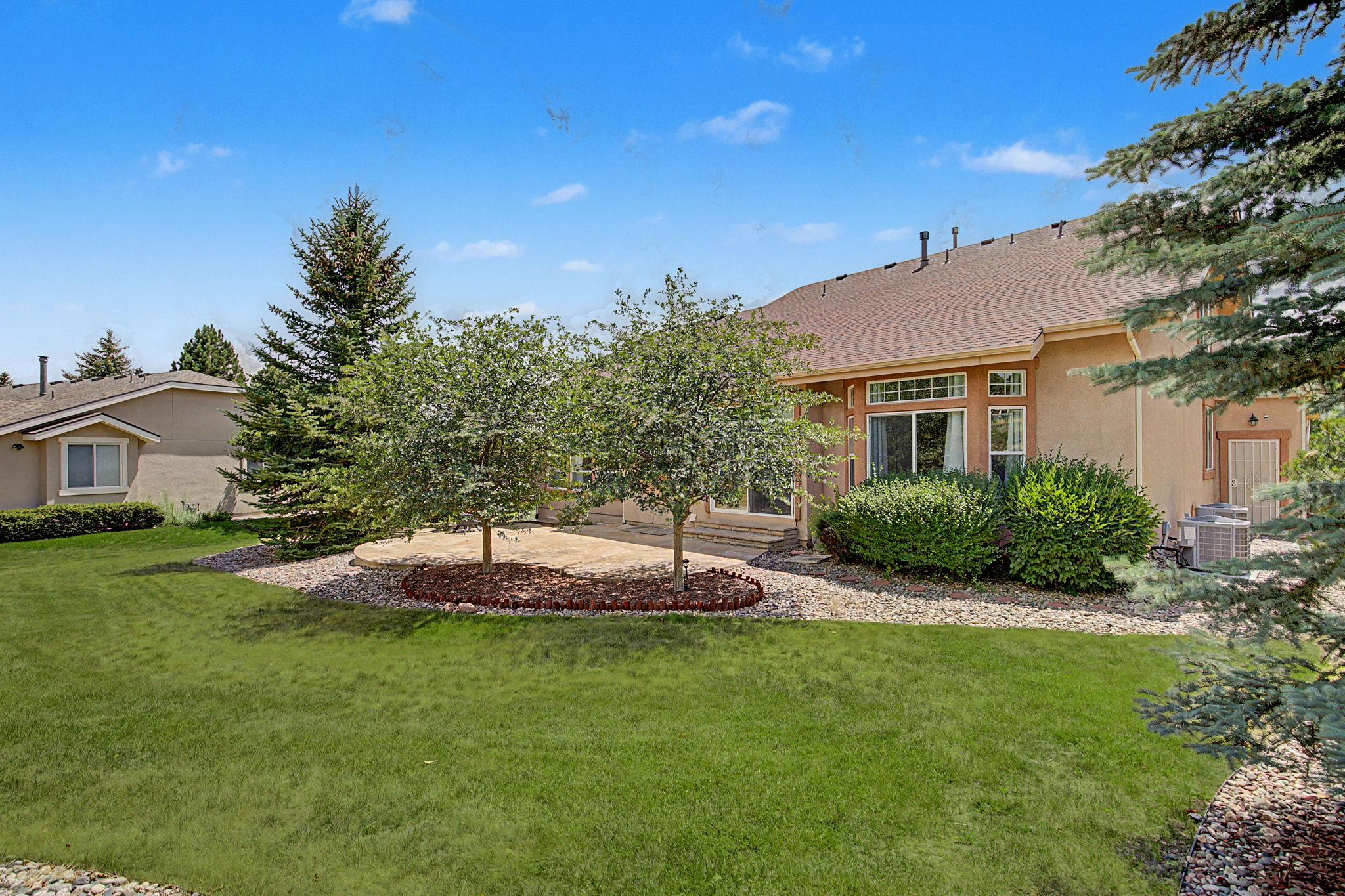 This rear view of a suburban home showcases a spacious backyard with a well-maintained lawn and a stone patio area. Two mature trees provide shade over the patio, which is bordered by a decorative stone edge, while large windows and a sliding glass door offer a clear connection to the interior living space. The scene is framed by evergreen trees and a clear blue sky, creating a peaceful and inviting outdoor atmosphere.