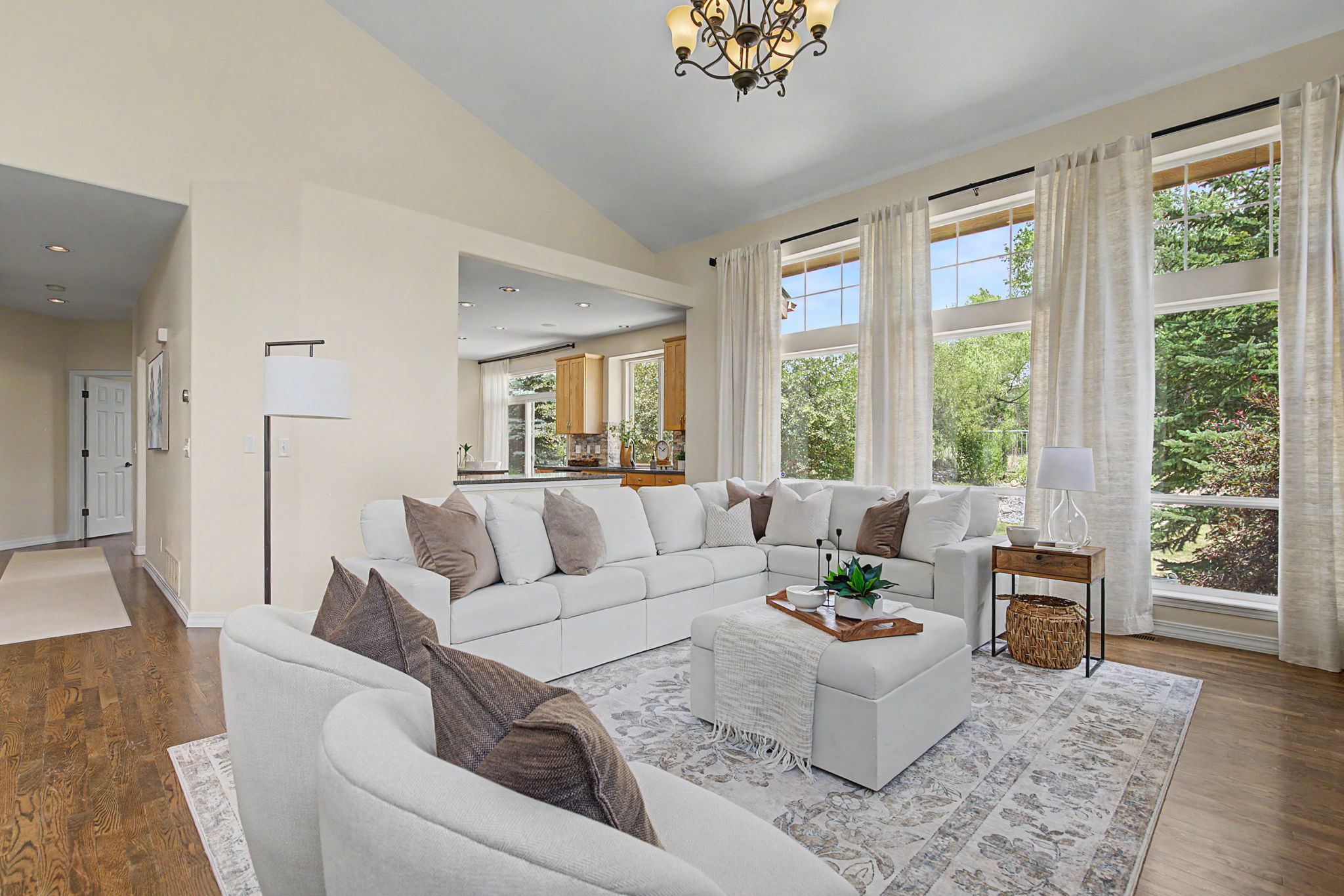 This bright and airy living room features a large, neutral-toned sectional sofa arranged around a central ottoman, creating a comfortable gathering space. Tall windows with light curtains allow ample natural light to fill the room, highlighting the vaulted ceiling and the open-concept layout that flows into the adjacent kitchen. The space is accented by a patterned area rug, a decorative chandelier, and a floor lamp, offering a warm and inviting atmosphere.