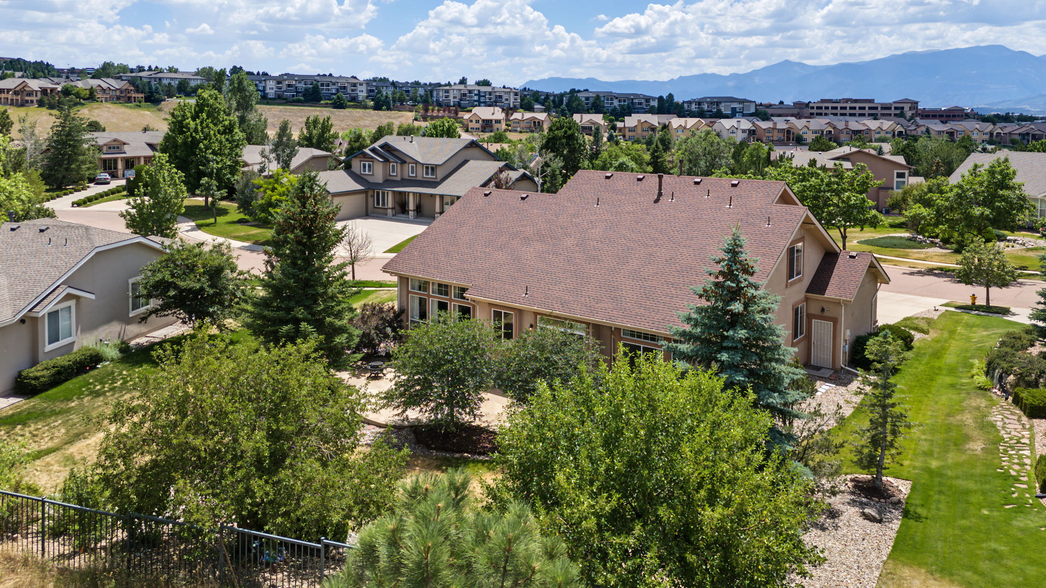 This high-angle aerial view captures a suburban residential property featuring a large, multi-gabled home with a brown shingled roof. The backyard includes a paved patio area surrounded by mature trees and landscaping, transitioning into a well-maintained lawn. In the background, a sprawling neighborhood is visible under a bright, cloudy sky with distant mountains on the horizon.