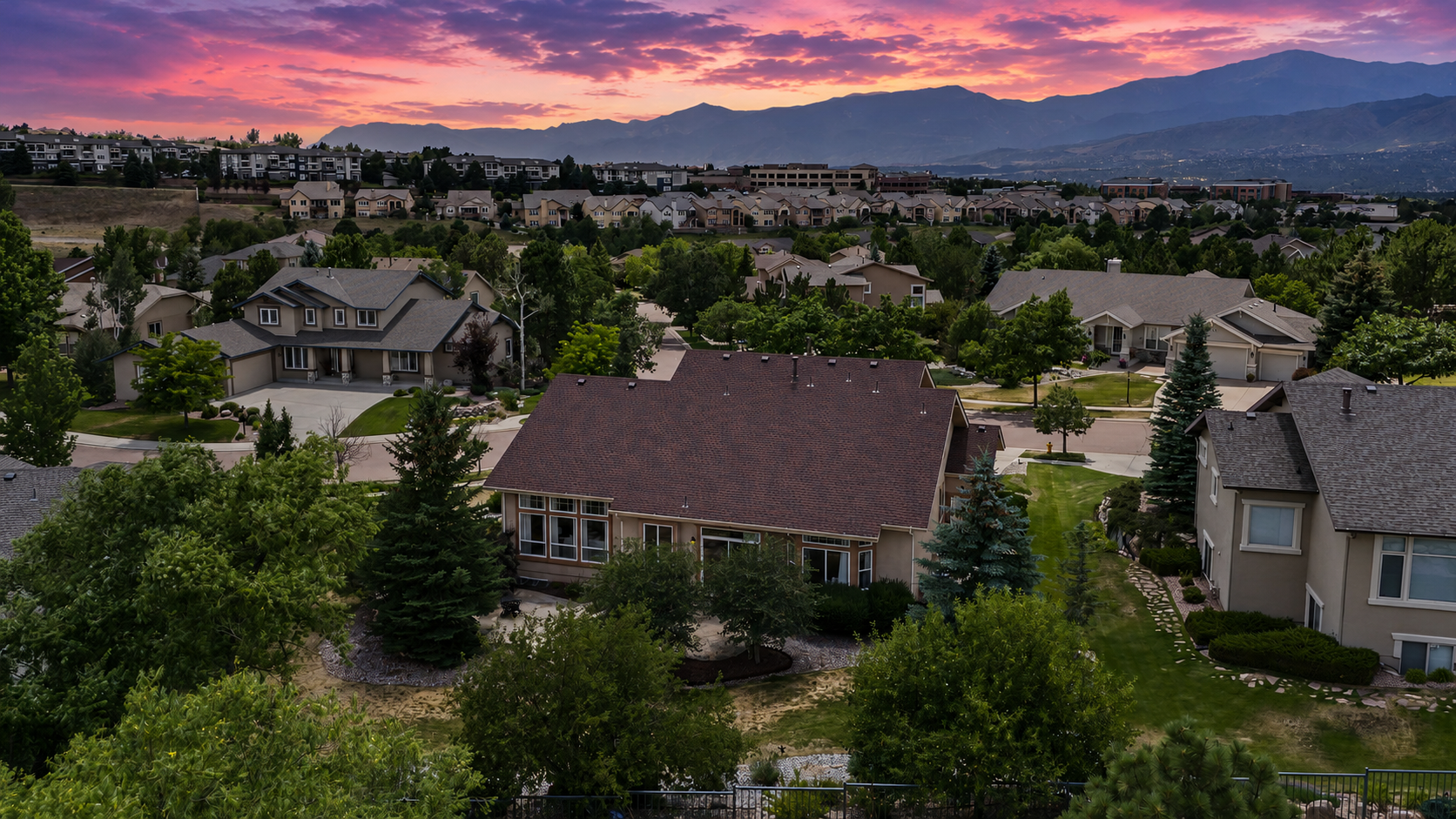 This high-angle aerial view captures a suburban neighborhood at sunset, showcasing a large residential property with a spacious backyard and patio area. The scene features a mix of single-family homes and multi-unit dwellings nestled among mature trees, with a dramatic, vibrant mountain range silhouetted against a colorful, cloud-filled sky. The perspective emphasizes the expansive layout of the community and the peaceful, scenic setting of the home.