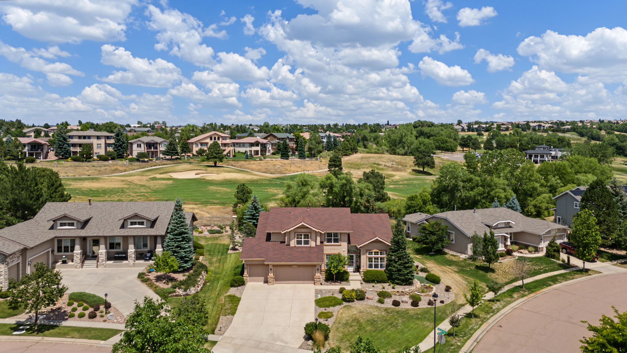 This high-angle aerial view captures a suburban neighborhood featuring several single-family homes with brown shingled roofs and neutral-toned siding. The properties are situated along a curved street, with a sprawling, grassy golf course visible in the background under a bright blue sky filled with scattered clouds. The scene emphasizes the spacious layout of the community, showcasing well-maintained lawns, mature trees, and a peaceful residential atmosphere.