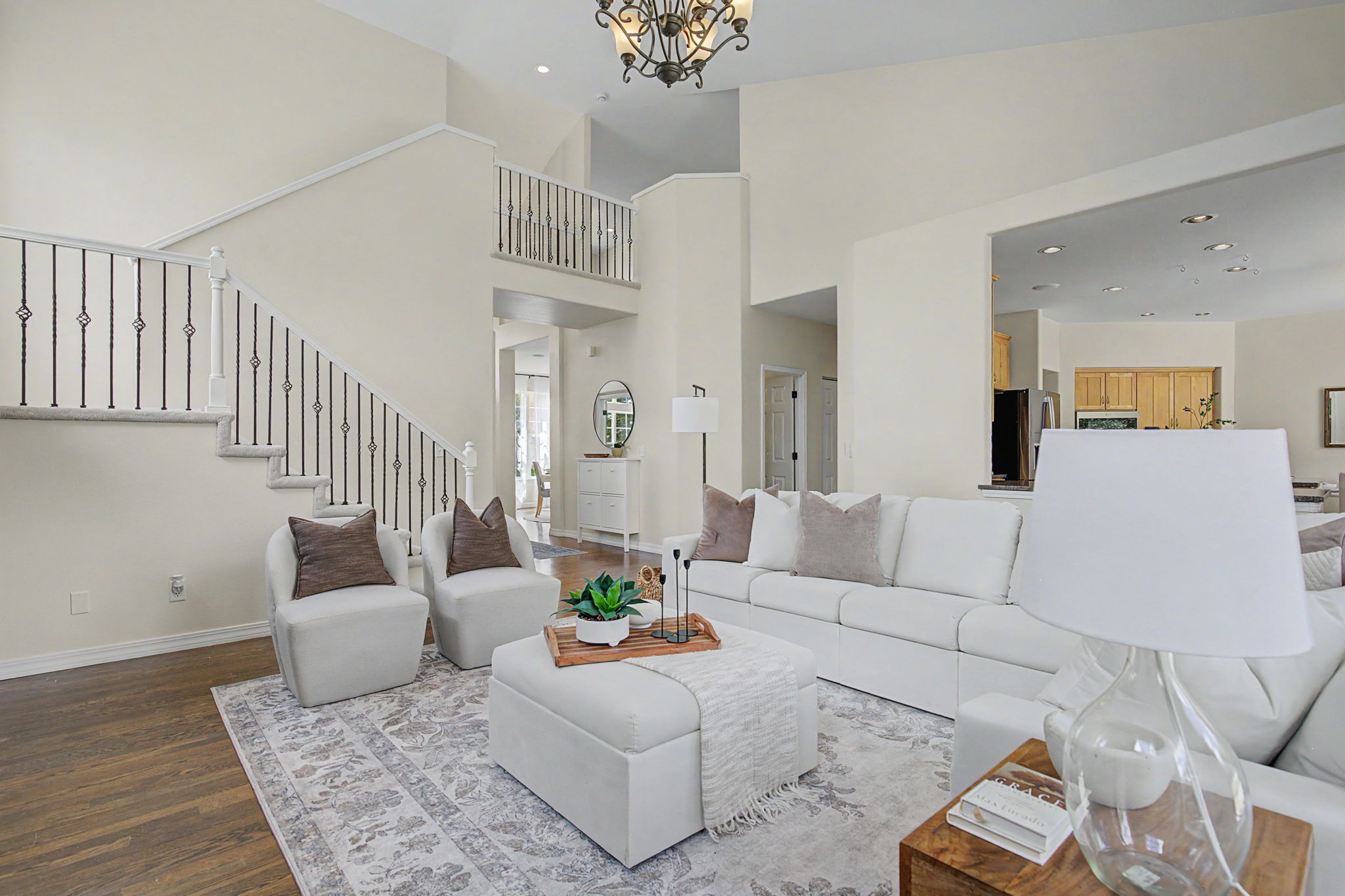 This bright and airy living room features a high, vaulted ceiling and an open-concept layout that flows into the kitchen area. The space is furnished with a large white sectional sofa, a matching ottoman, and two accent chairs, all arranged on a patterned area rug over dark wood flooring. A staircase with decorative metal railings leads to an upper landing, creating a sense of architectural depth and openness.