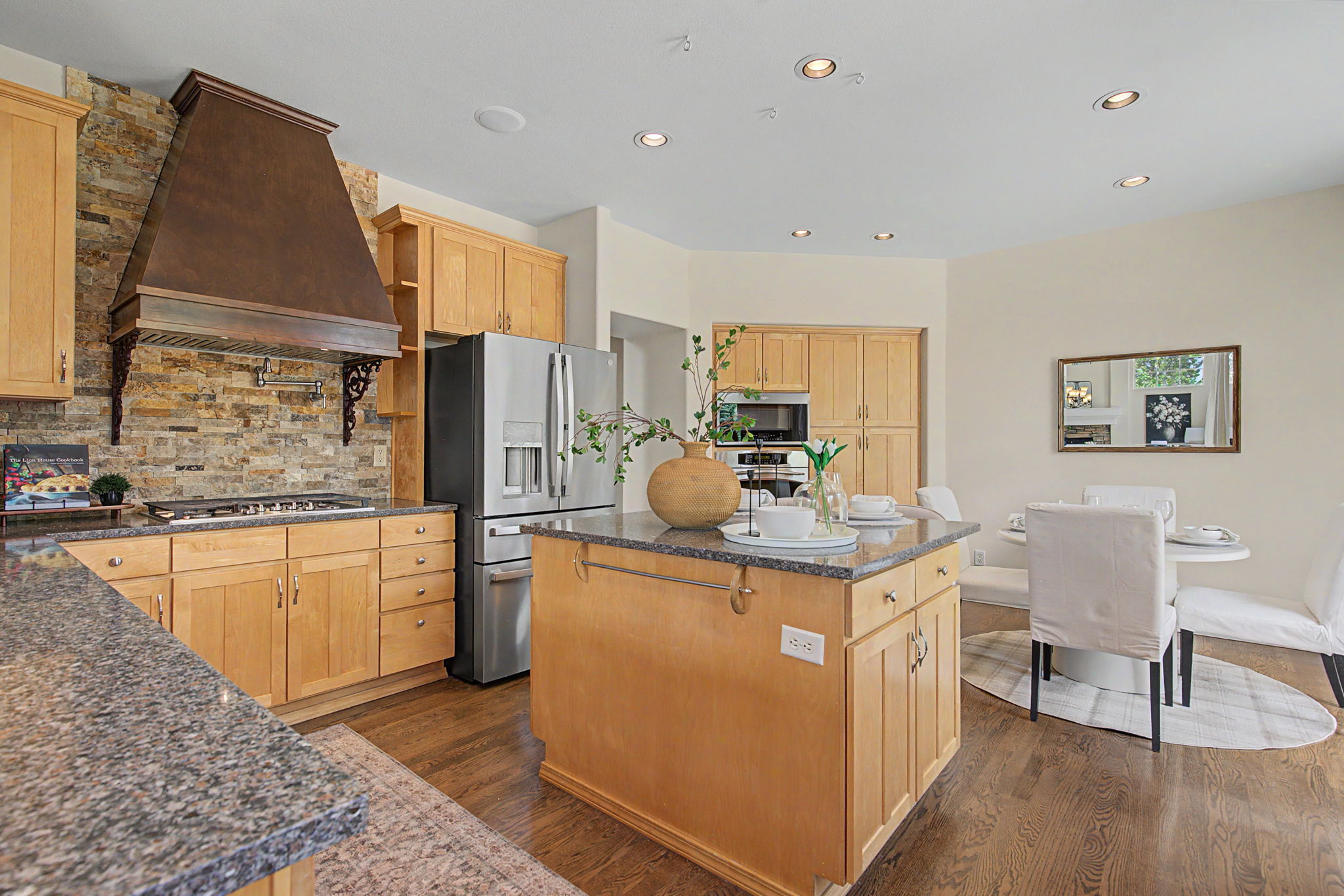This spacious kitchen features light wood cabinetry, dark stone countertops, and a central island with integrated seating. A prominent stone-clad chimney breast houses the range and a large custom hood, creating a rustic focal point. The open-concept layout flows seamlessly into a bright dining area, making it an ideal space for both cooking and entertaining.