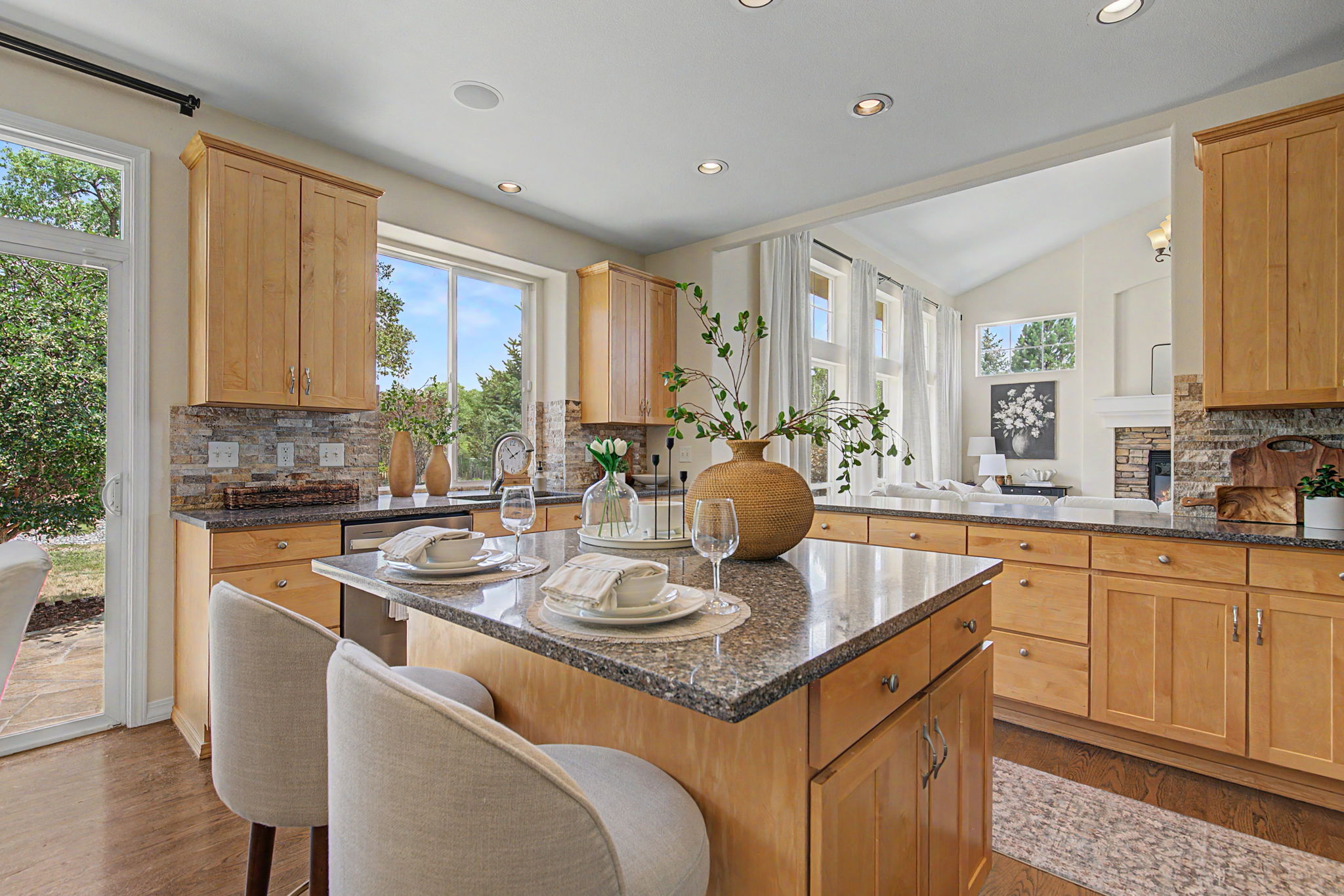 This bright, open-concept kitchen features light wood cabinetry, stone countertops, and a central island with bar seating. The space is well-lit by recessed lighting and natural light from a glass door and a large window, creating an inviting atmosphere. Beyond the kitchen island, a glimpse into the adjacent living area reveals a stone-clad chimney breast, emphasizing the home's cohesive and warm design.