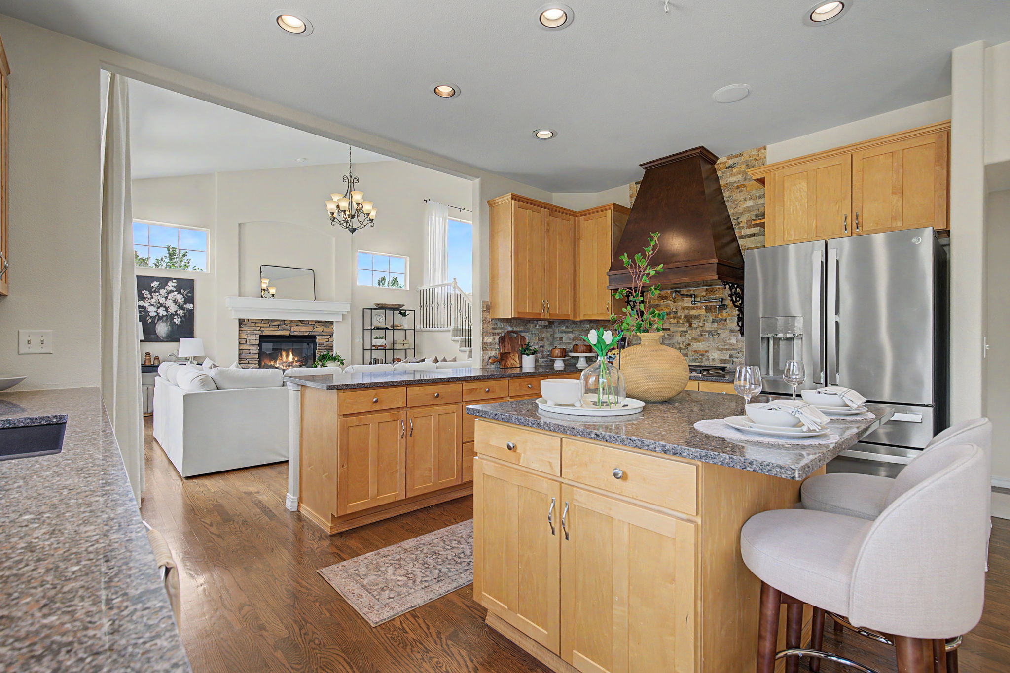 This open-concept kitchen features light wood cabinetry, stone countertops, and a central island with bar seating. A prominent dark wood range hood sits above the stove, complemented by a stone-clad backsplash, while the space flows seamlessly into a living area with a stone fireplace. The perspective captures the functional kitchen workspace in the foreground, looking through to the bright, airy living room beyond.