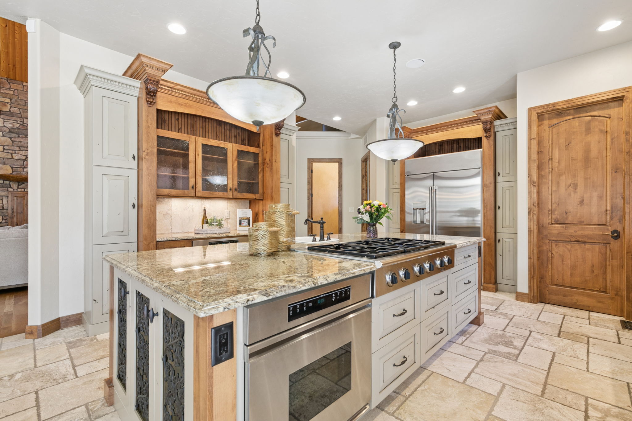 This spacious kitchen features a large central island with a stone countertop, a built-in gas cooktop, and an integrated oven. The cabinetry combines light-toned painted wood with natural wood accents, complemented by decorative metal panels on the island and two ornate pendant lights overhead. The room is finished with light-colored stone tile flooring and includes a stainless steel refrigerator, creating a warm and traditional aesthetic.