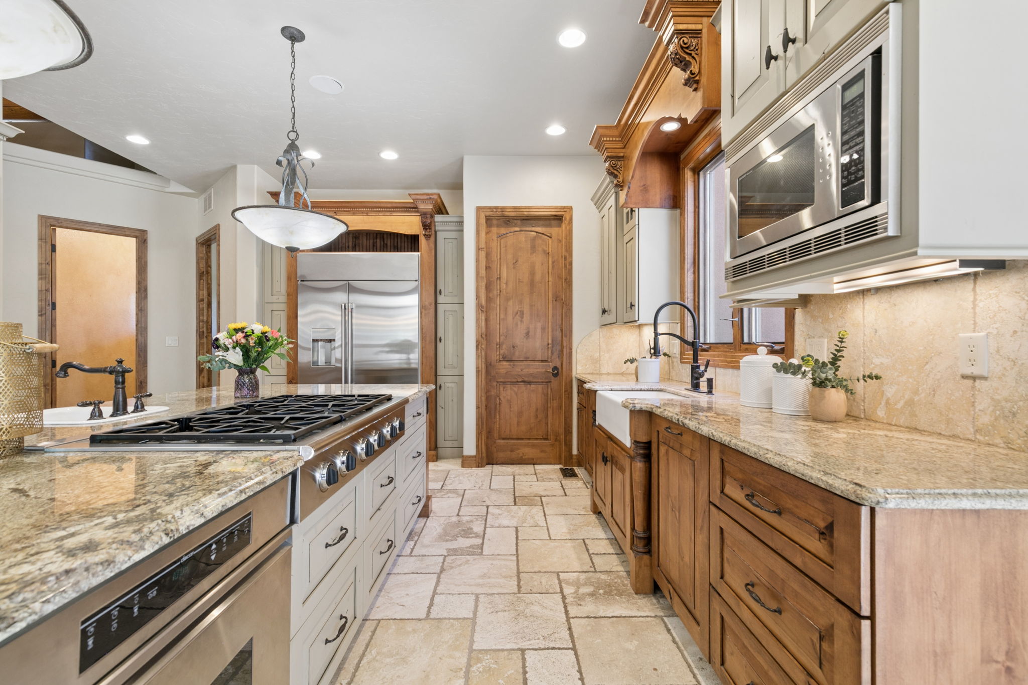 This spacious kitchen features a blend of light-toned cabinetry on the island and rich, warm-toned wood cabinetry along the perimeter. A large central island houses a gas cooktop and provides ample counter space, complemented by stone countertops and a farmhouse-style sink. The room is finished with patterned stone flooring and decorative woodwork, creating a traditional and inviting atmosphere.