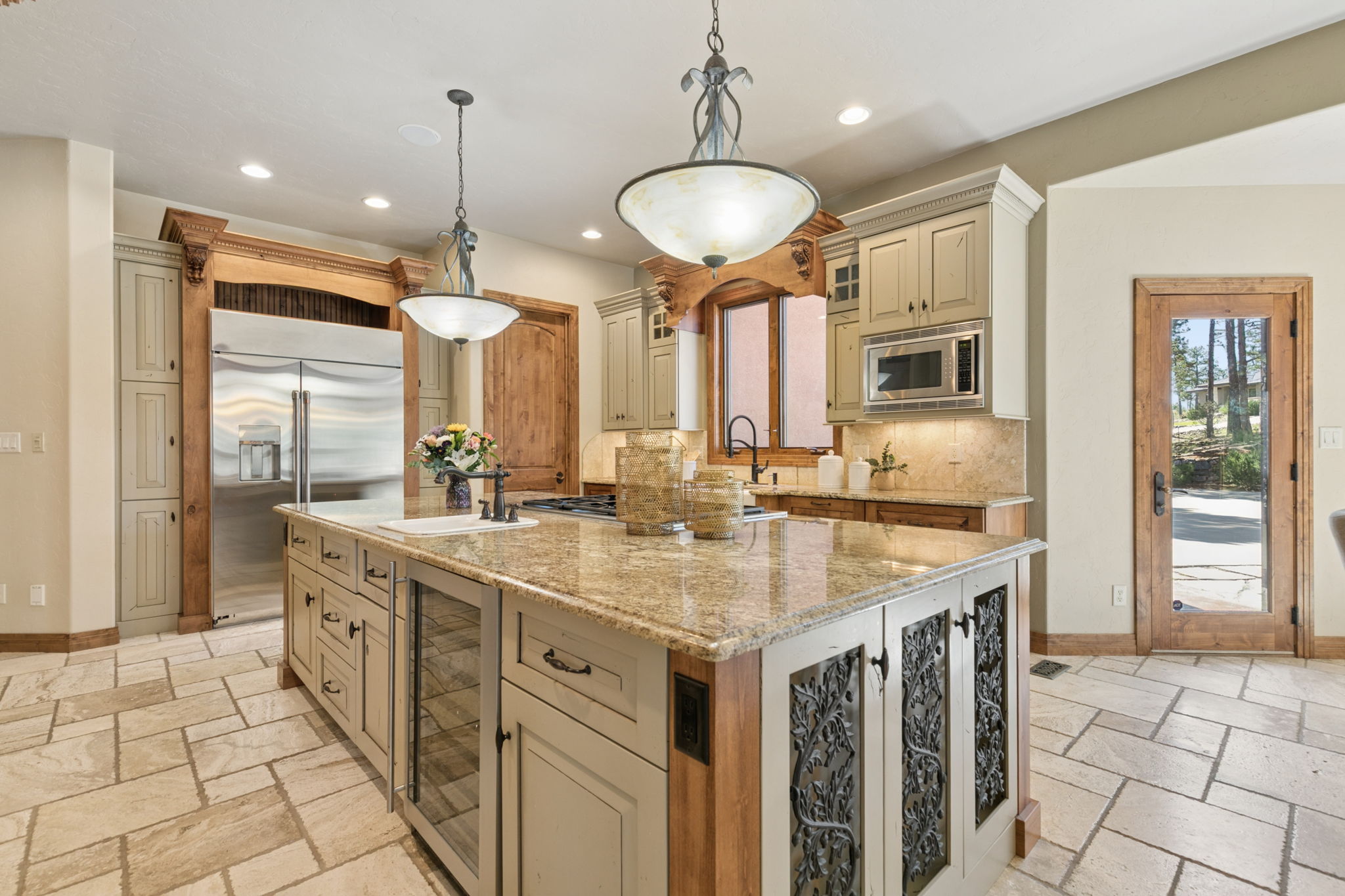 This spacious kitchen features a large central island with a stone countertop, integrated wine storage, and decorative metal-grille cabinet doors. The room is anchored by light-toned cabinetry, stainless steel appliances, and warm wood accents, including a custom hood surround above the cooktop. Two elegant pendant lights hang over the island, while a glass-paned door provides a view of the outdoor landscape, creating a bright and inviting atmosphere.