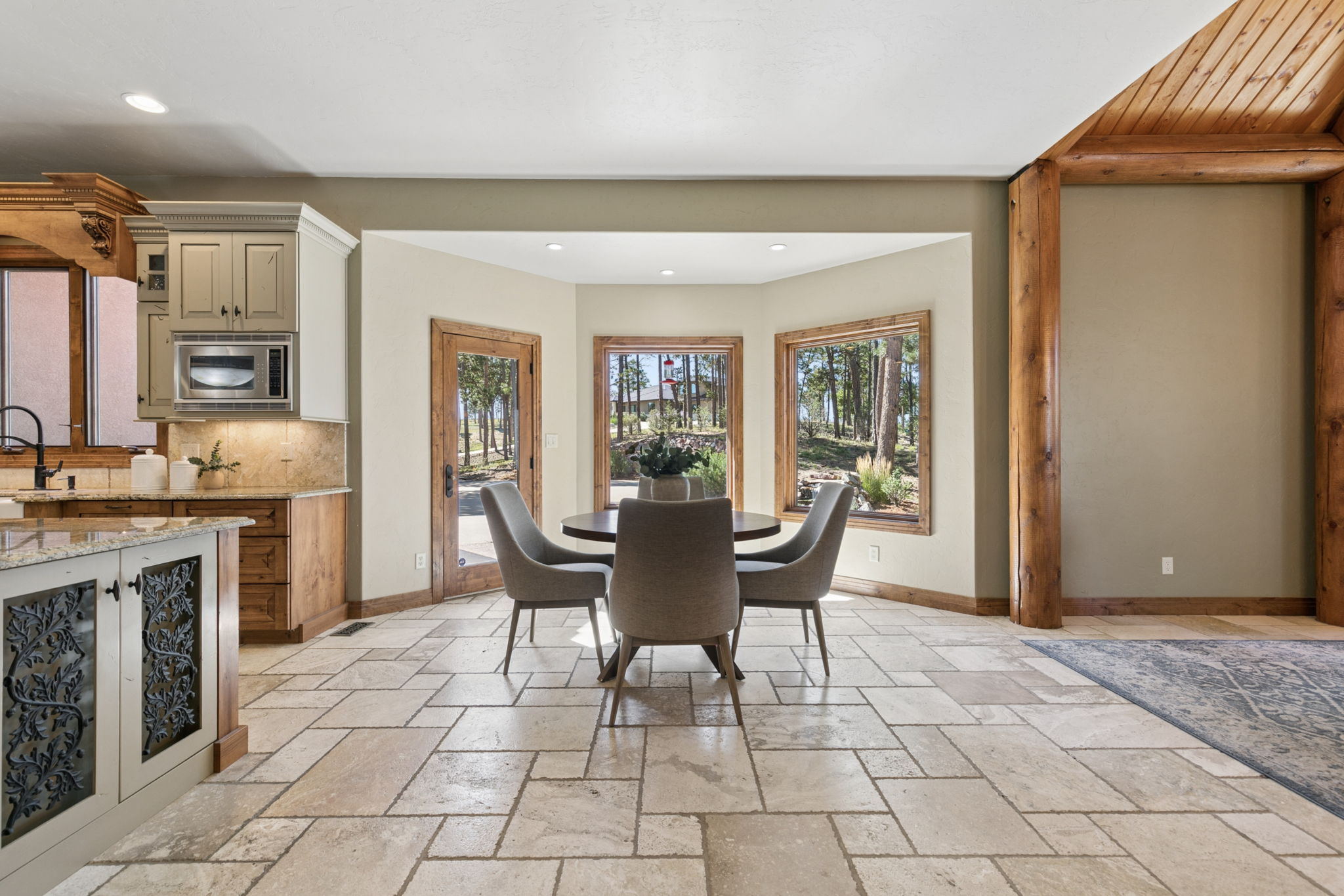 This bright kitchen and dining area features a neutral color palette with light-toned cabinetry and a stone-topped island with decorative metal detailing. A circular dining table with four upholstered chairs sits in a sunlit bay window nook, offering views of the surrounding forest. The space is anchored by large-format stone tile flooring and a prominent natural wood support post, creating a warm, rustic-inspired atmosphere.