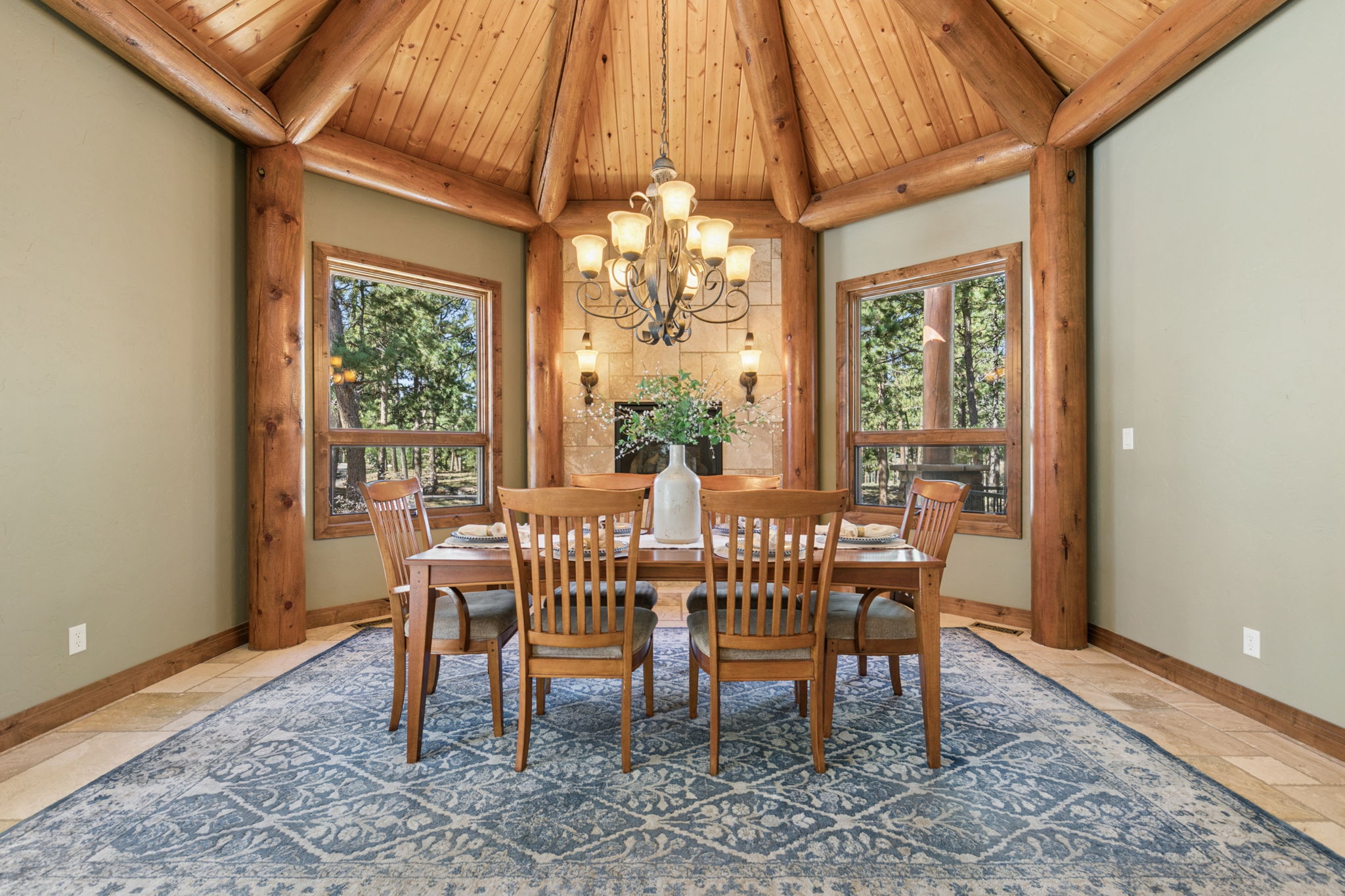 This rustic dining room features a central wooden table surrounded by six chairs, set atop a large, intricate blue patterned area rug. The space is defined by a vaulted, wood-paneled ceiling with exposed log beams and a stone-clad chimney breast that serves as a focal point behind the table. Large windows on either side of the fireplace provide ample natural light and views of the surrounding trees, creating a warm and inviting atmosphere.