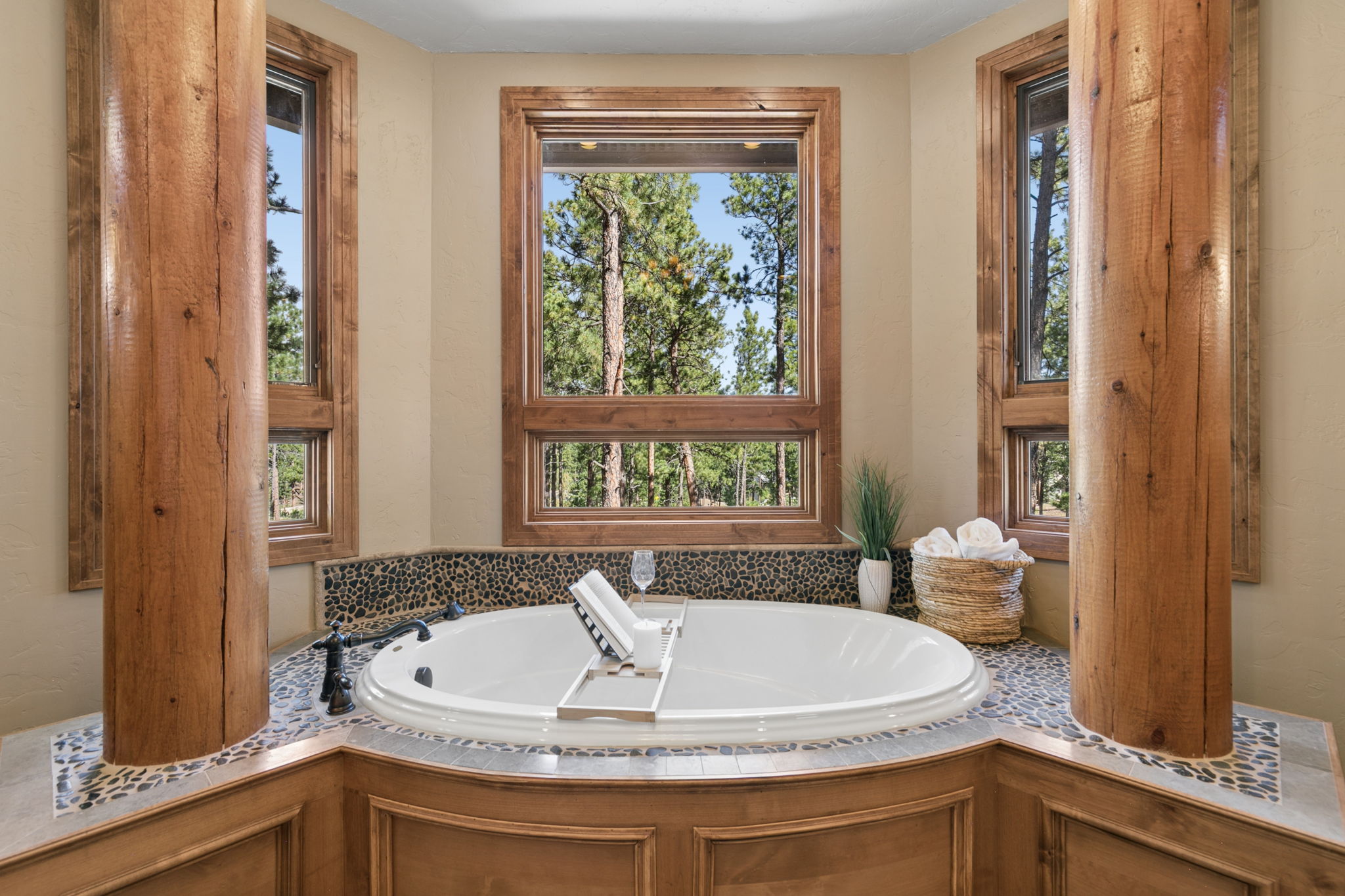 This primary bathroom features a large, oval soaking tub nestled into a custom alcove framed by two prominent, rustic log pillars. The tub surround is finished with decorative pebble-style tile, and a wooden tray holds a book and a glass, while a nearby basket contains rolled towels. Large windows behind the tub provide a serene, elevated view of the surrounding forest, creating a tranquil and spa-like atmosphere.