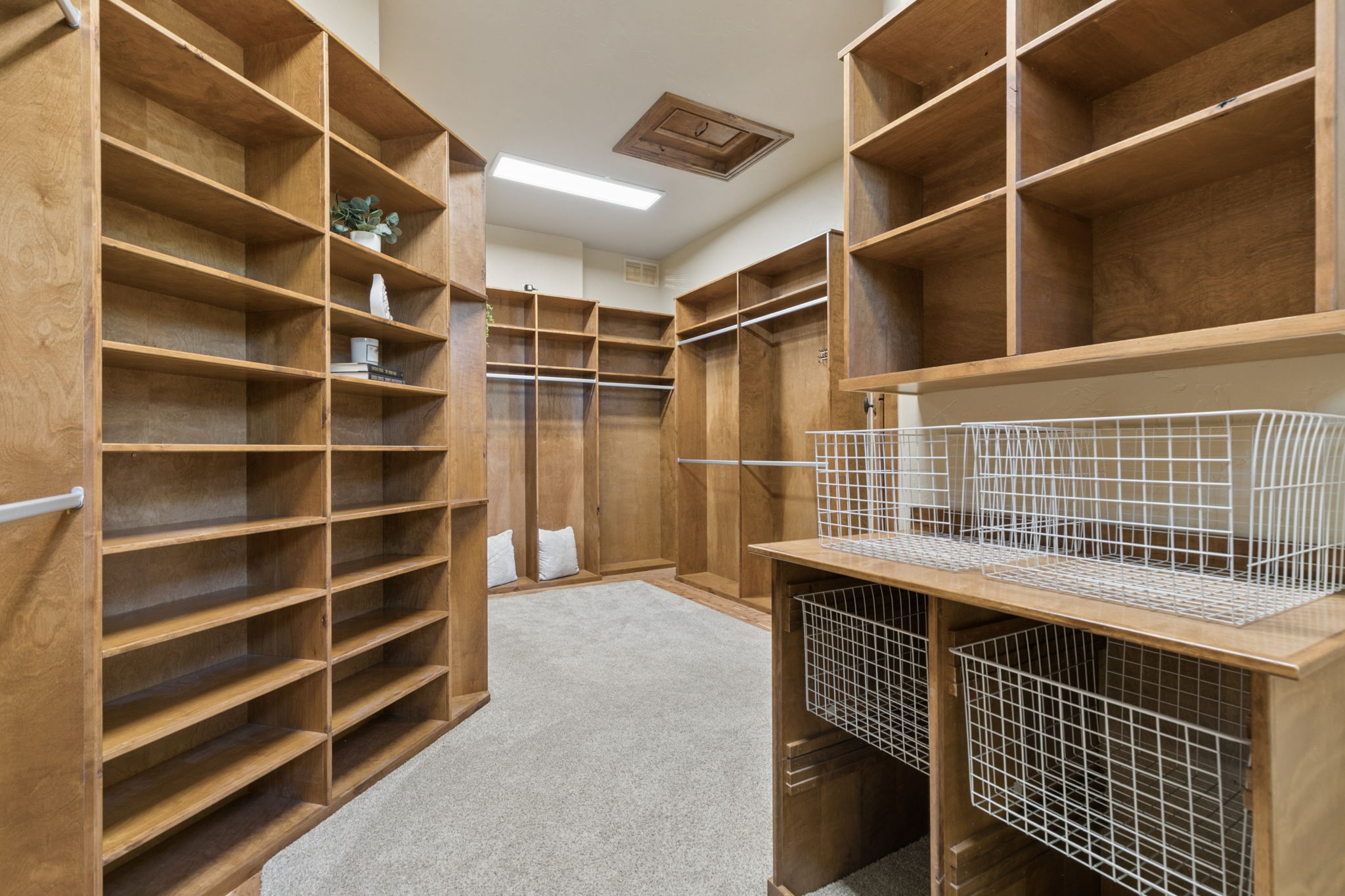 This spacious walk-in closet features extensive custom wood shelving and hanging rods, providing ample storage and organization options. The room is carpeted and includes a built-in workstation area with wire baskets, offering a functional layout for both clothing and accessories. The perspective is from the entrance, looking into the deep, well-lit space that maximizes every inch of wall area.