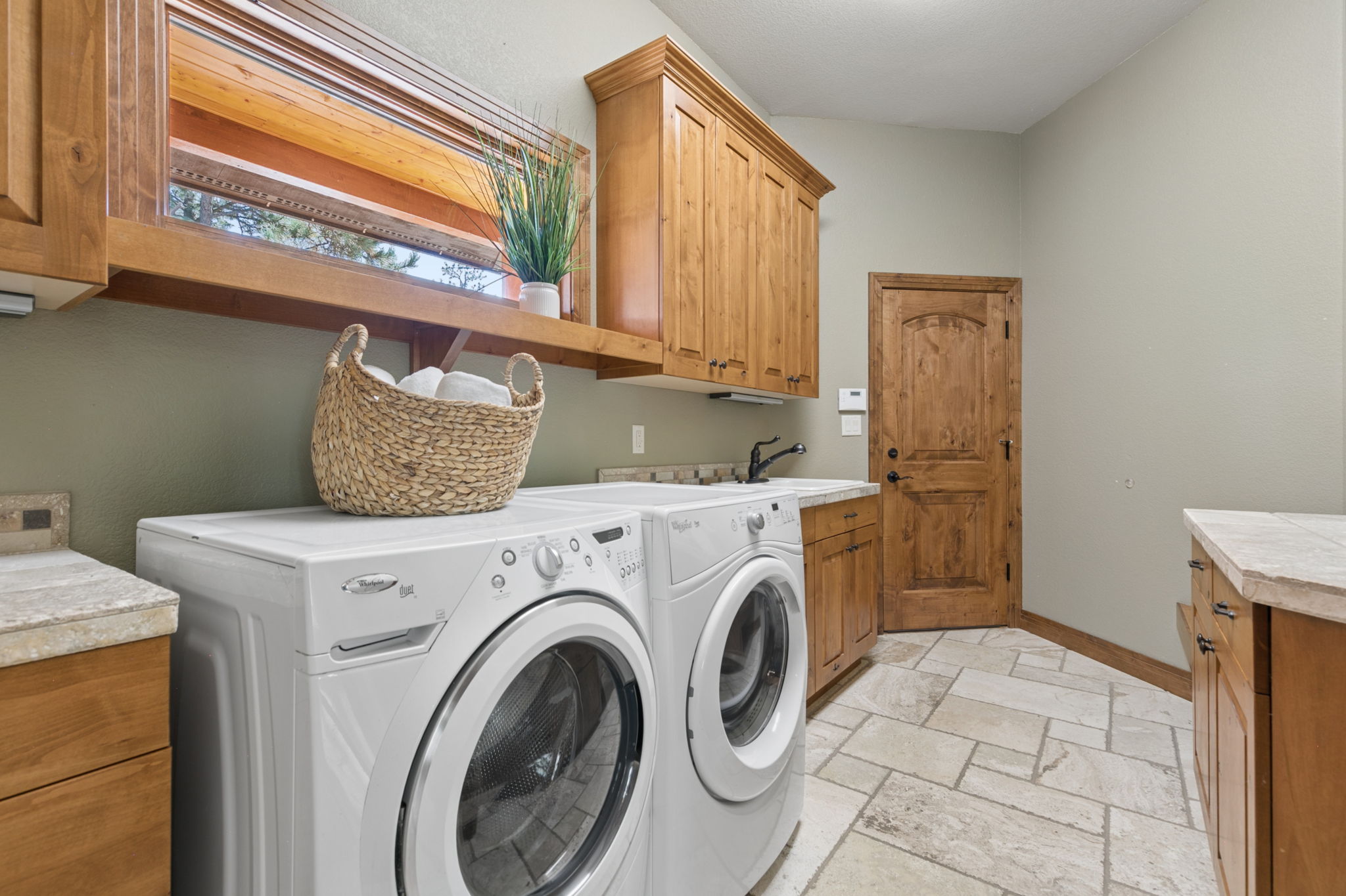 This functional laundry room features a pair of white front-loading appliances positioned beneath a wooden shelf and a set of upper cabinets. The space includes a utility sink, stone-topped counters, and light-colored tile flooring, all complemented by a warm-toned wooden door. The room is well-lit and maintains a cohesive, rustic-inspired aesthetic.