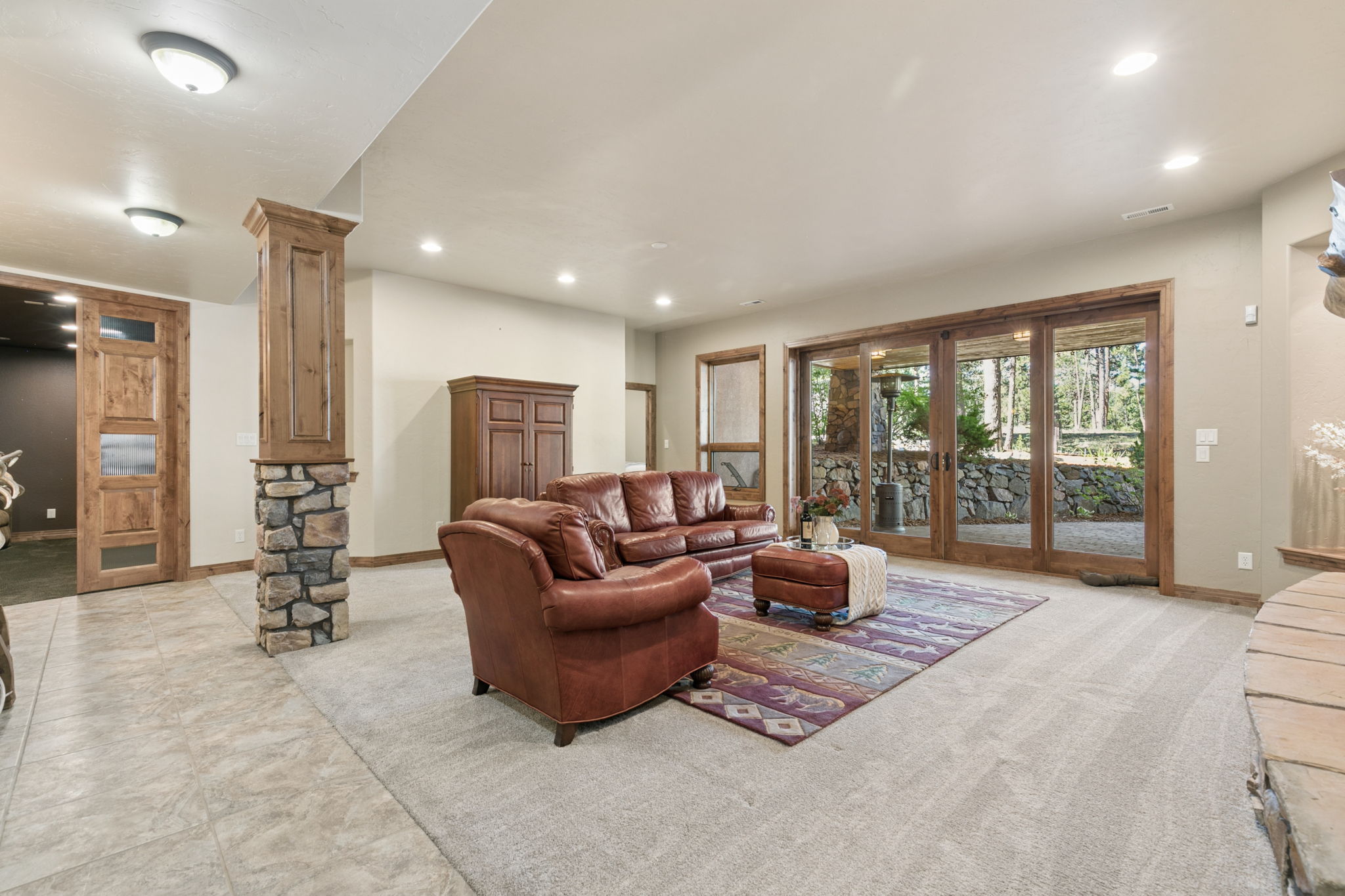 This spacious living area features a warm, rustic aesthetic with a leather sofa and armchair set arranged on a patterned area rug. A prominent stone-clad pillar anchors the room, while large glass sliding doors provide a clear view of the wooded outdoor patio area. The neutral walls and carpeted flooring create a comfortable, inviting atmosphere suitable for relaxation.