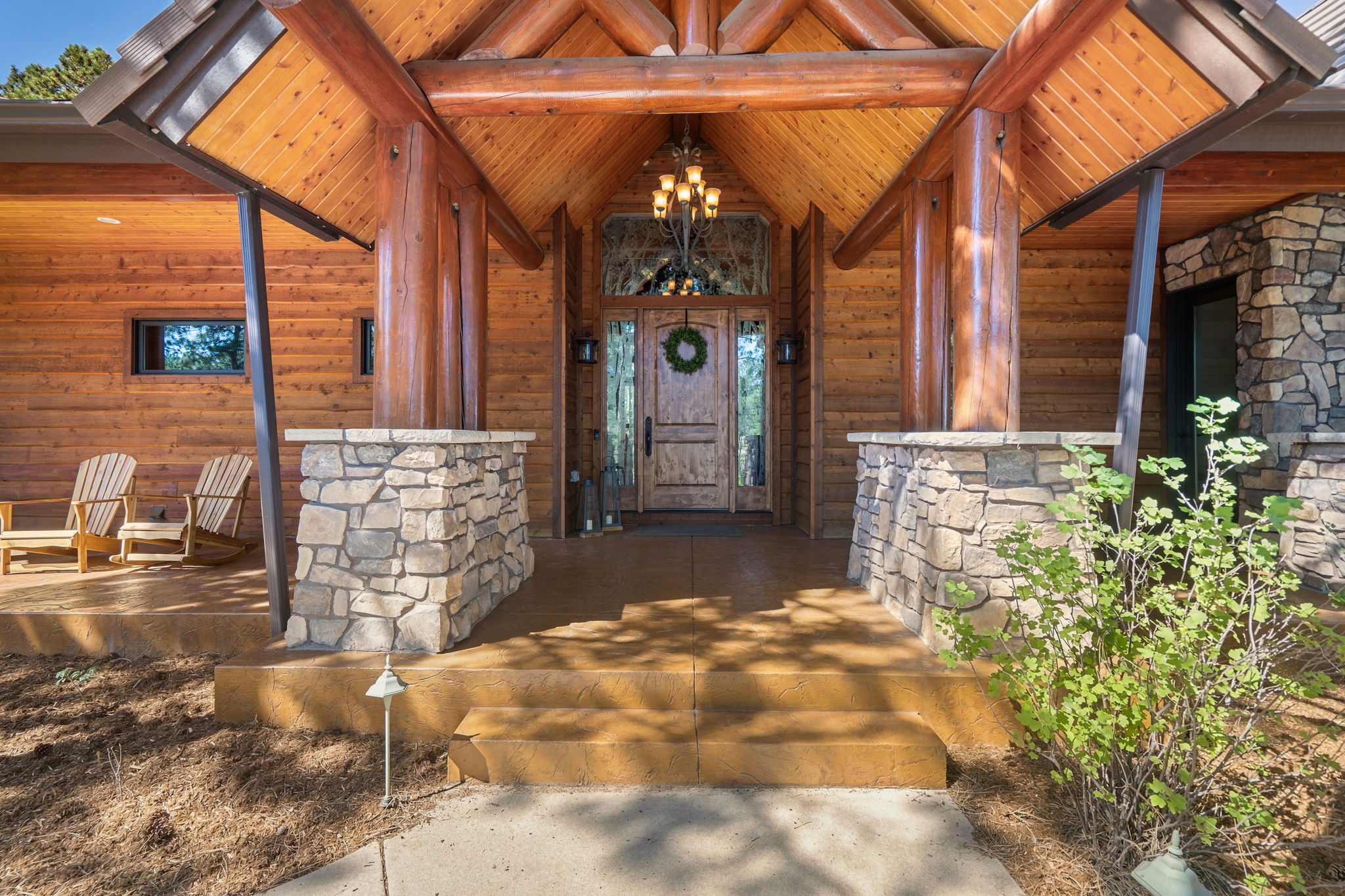 This inviting log cabin-style entryway features a prominent covered porch supported by thick, rustic timber columns resting on stone-clad bases. The front door is framed by a decorative transom window and flanked by matching wall sconces, while a pair of wooden rocking chairs sits on the left side of the porch. The scene is captured from a low, centered perspective, emphasizing the warm, natural wood textures and the welcoming, elevated entrance.