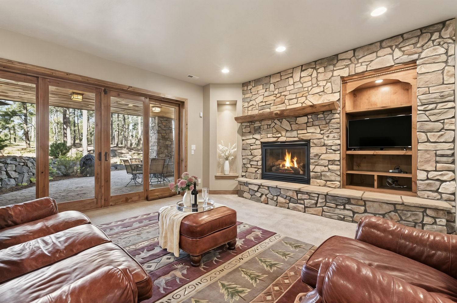 This cozy living room features a prominent stone-clad chimney breast with a built-in fireplace and a rustic wood mantel. To the right, a custom wood shelving unit houses a television, while a large set of sliding glass doors opens to a covered patio, providing a seamless connection to the wooded outdoors. The space is furnished with warm leather seating and a patterned area rug, creating a comfortable and inviting atmosphere.