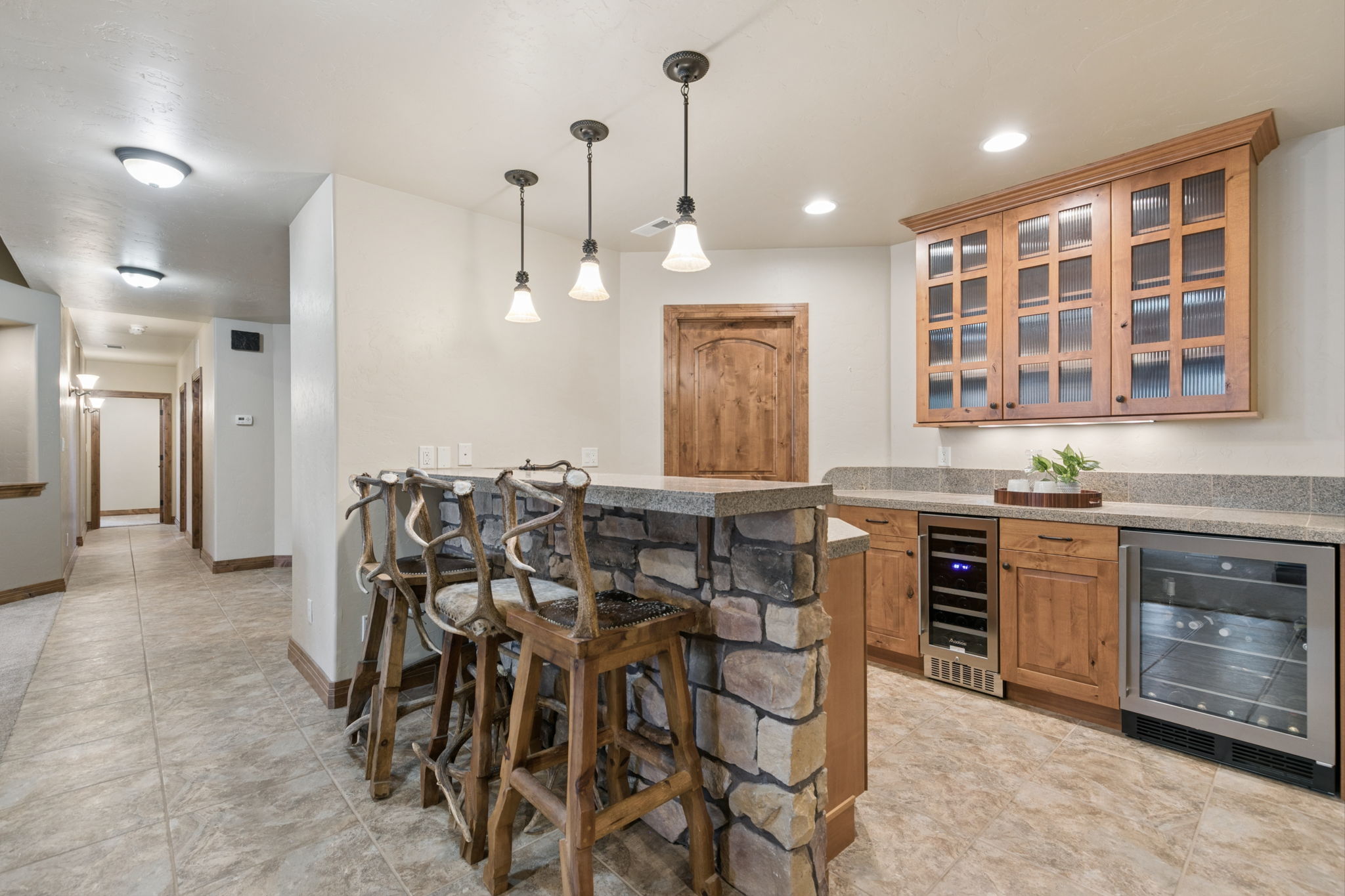 This rustic-style wet bar area features a stone-clad base with a countertop and three unique bar stools crafted from natural wood and antlers. The space includes a built-in wine refrigerator, a beverage cooler, and upper cabinetry with glass-paneled doors, all set against a neutral wall backdrop. The perspective captures the bar from a slightly elevated angle, highlighting the transition from the tiled floor area into the adjacent hallway.