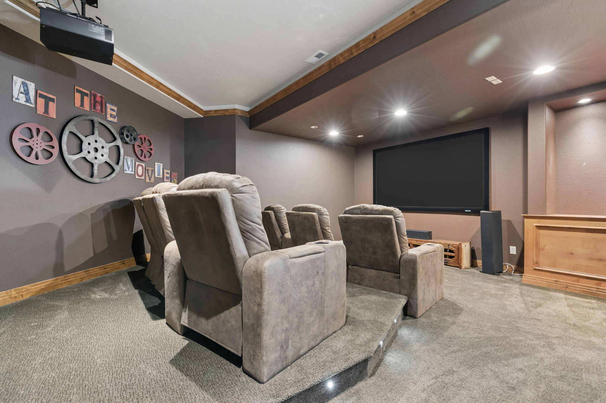 This dedicated home theater room features tiered seating with plush, brown reclining chairs arranged for optimal viewing of the large wall-mounted screen. The room is decorated with movie-themed wall art and includes recessed lighting, a projector mounted on the ceiling, and a wooden cabinet beneath the screen. The space is finished with dark-toned walls and carpeted flooring, creating an immersive cinematic environment.