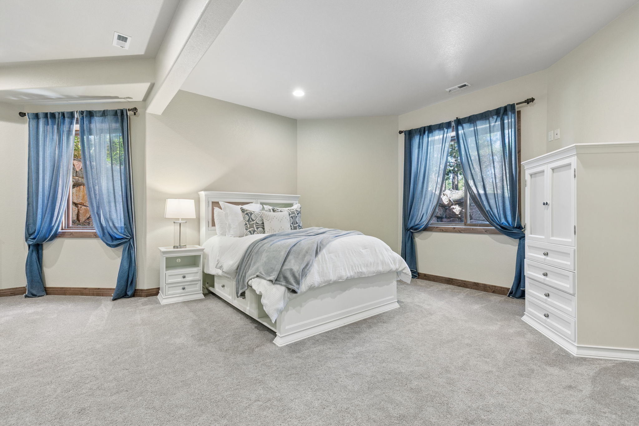This bright and spacious bedroom features neutral walls, plush carpeting, and a white bed frame centered between two windows draped in blue curtains. A matching white nightstand with a lamp sits to the left, while a tall white armoire provides storage on the right. The room offers a clean, serene atmosphere with natural light filtering through the windows.