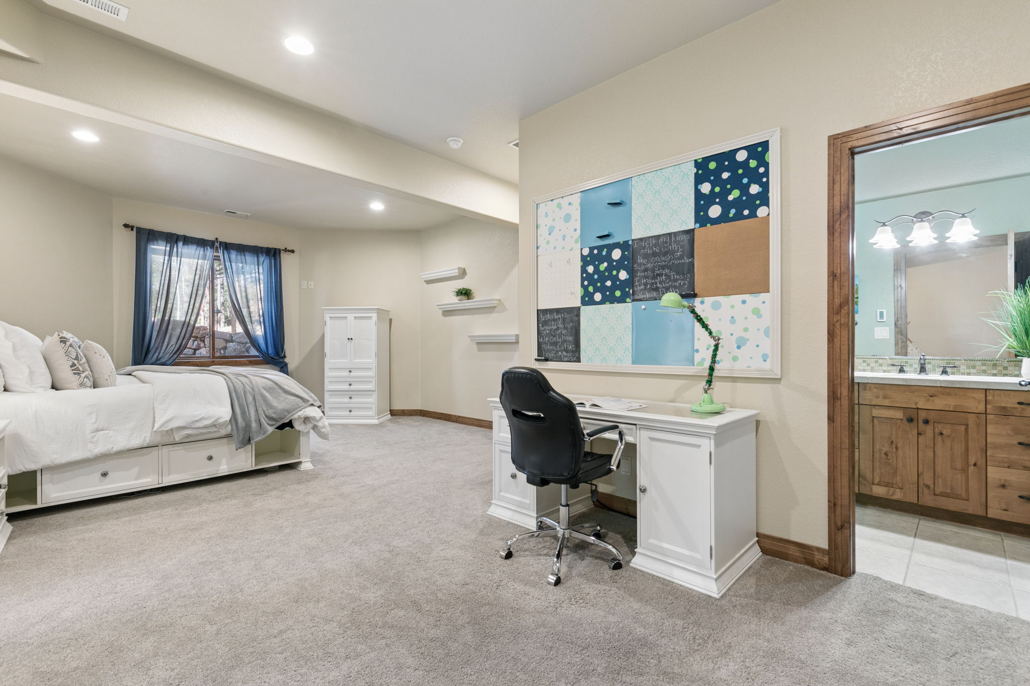 This spacious bedroom features a neutral color palette with plush carpeting and a white bed frame with built-in storage drawers. A dedicated study area is positioned against the wall, complete with a white desk, a black office chair, and a large, multi-paneled decorative bulletin board. To the right, an open doorway provides a view into an adjoining bathroom with a wood vanity and light-colored tile flooring.